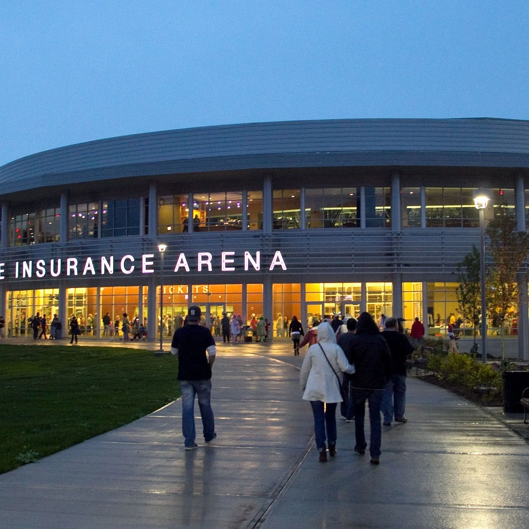 Exterior view of Erie Insurance Arena in Erie, Pennsylvania at dusk, featuring the illuminated entrance with crowds of people walking toward the venue, warm interior lighting visible through large windows, and a lively atmosphere as guests arrive for an event.