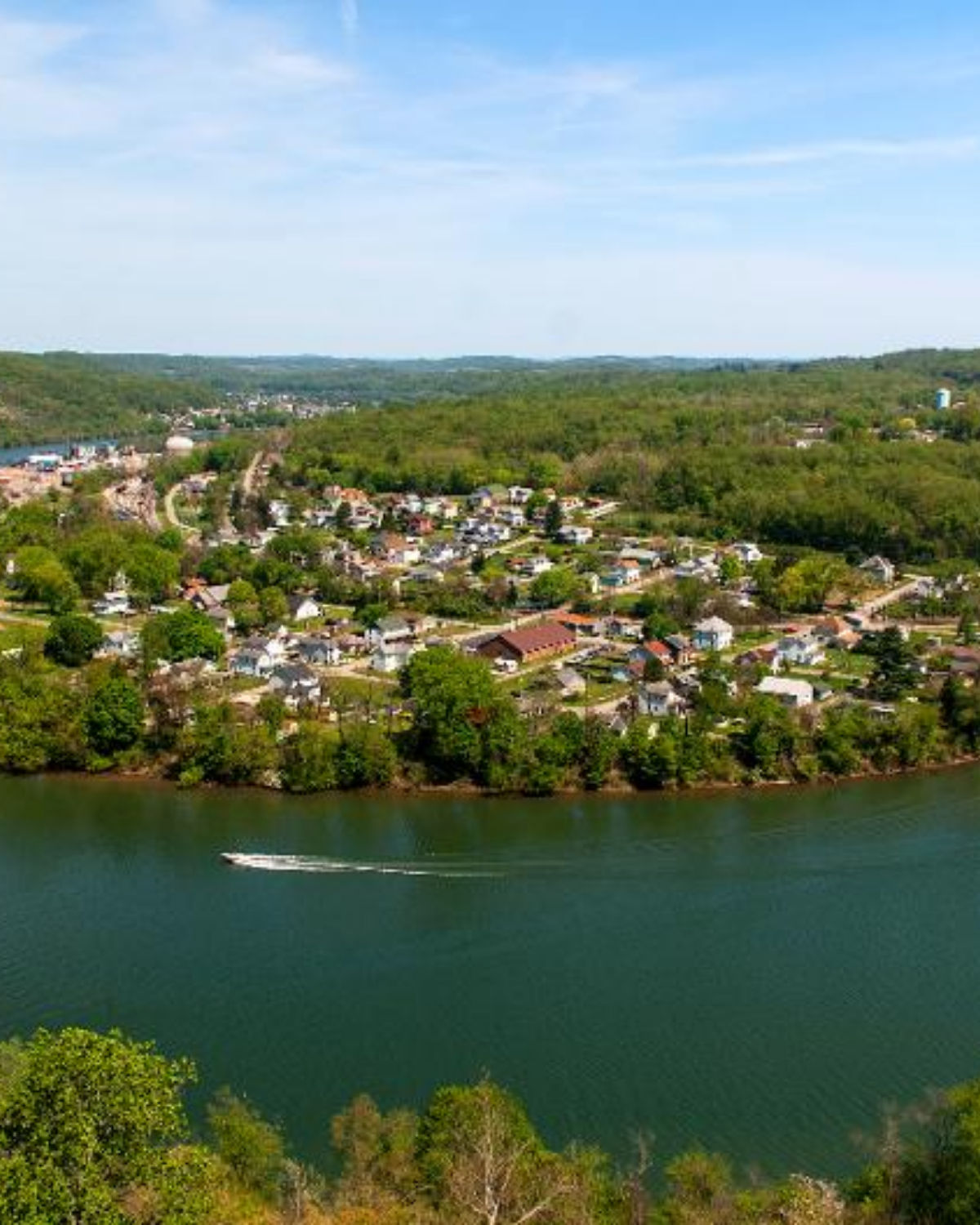 Aerial view of a small town with clustered houses and tree-covered hills along a river, with a boat leaving a white wake in the foreground.