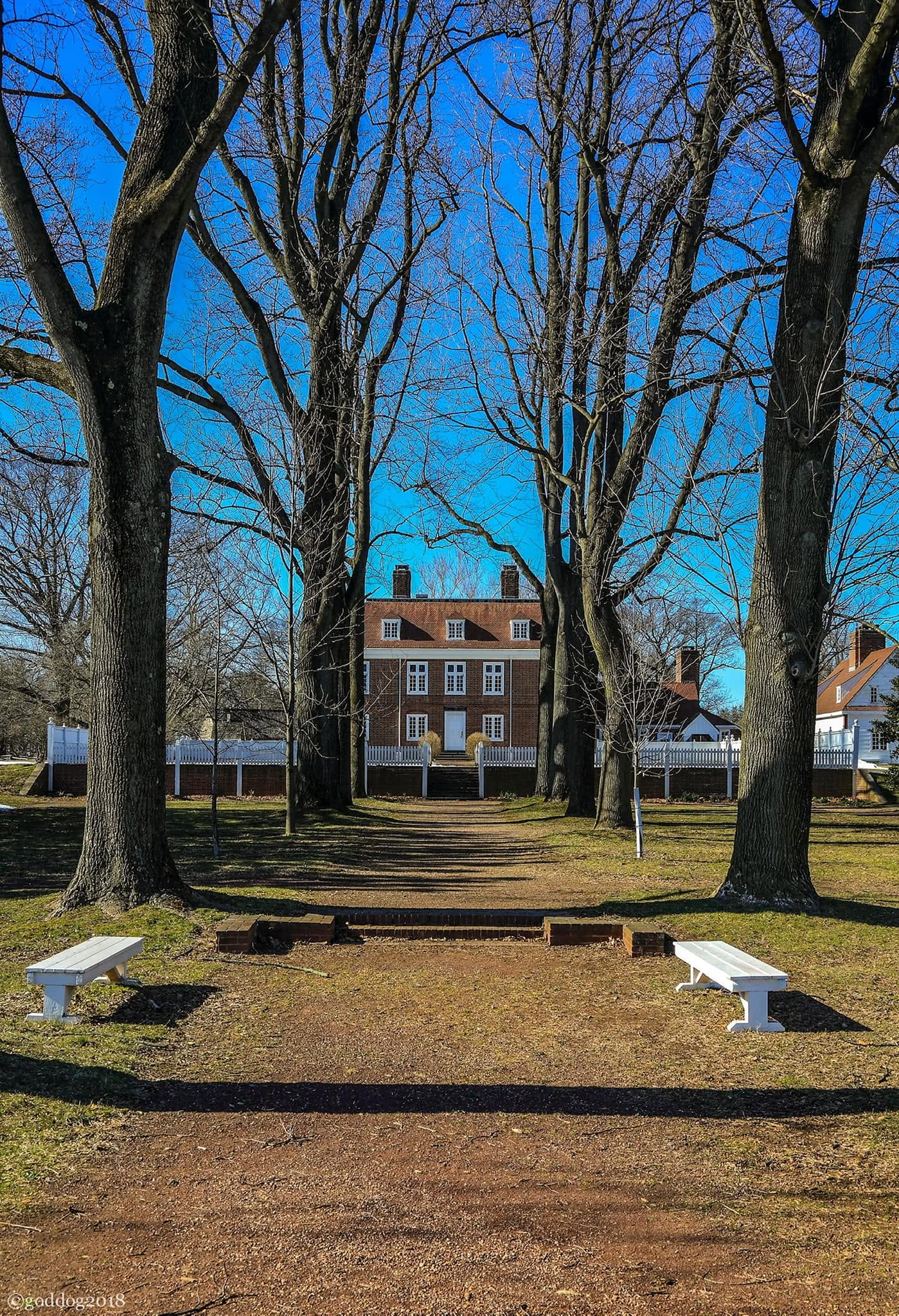 Pennsbury Manor in Levittown, Pennsylvania, viewed from a tree-lined pathway leading to the historic brick mansion. The preserved colonial estate features stately architecture, white fencing, towering trees, and landscaped grounds that reflect the area's rich Revolutionary-era history.