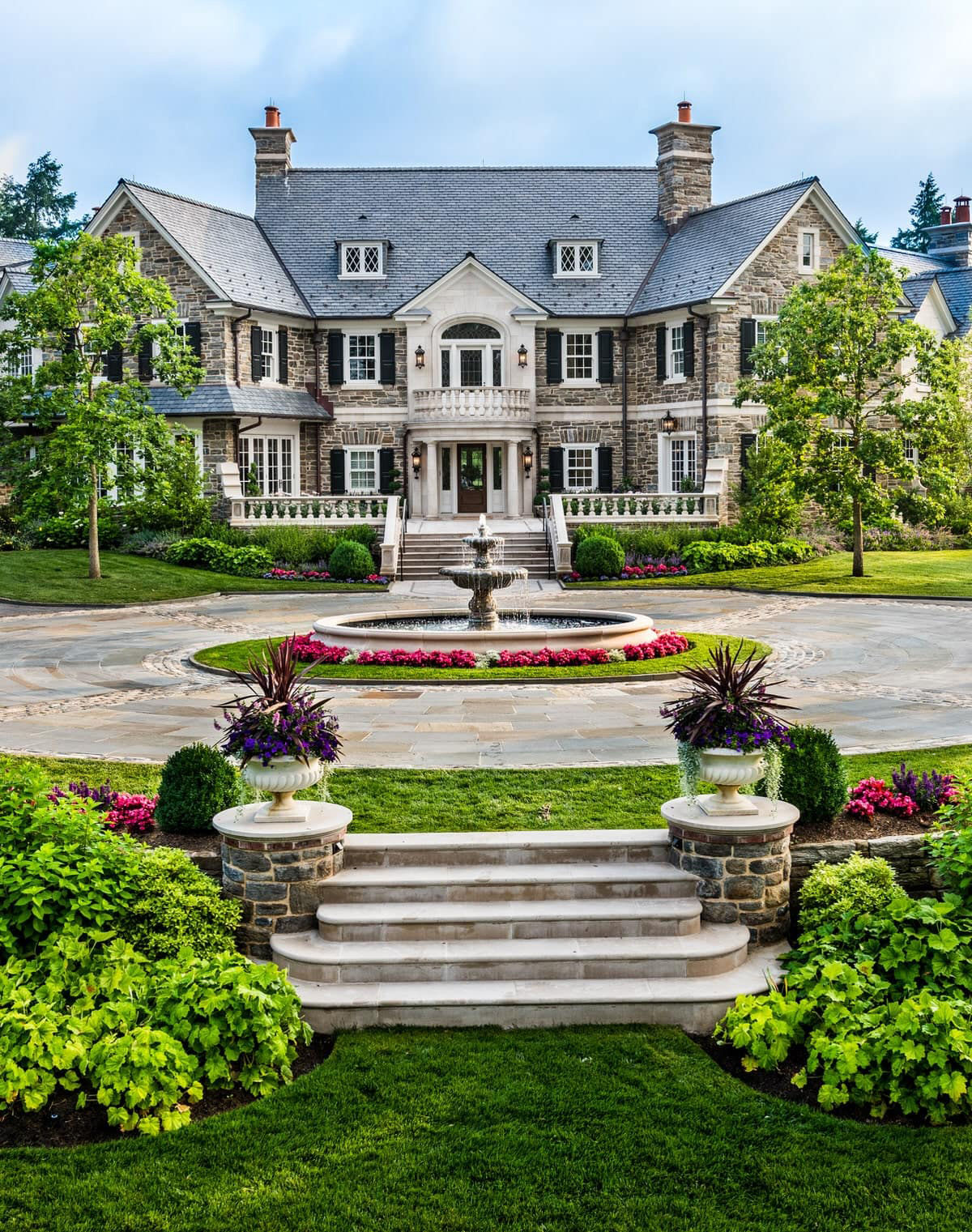 Front exterior of a stately stone estate in Merion Station, Pennsylvania, featuring classic architecture, manicured gardens, a circular driveway with a tiered fountain, symmetrical landscaping, and a grand entrance with stone steps.
