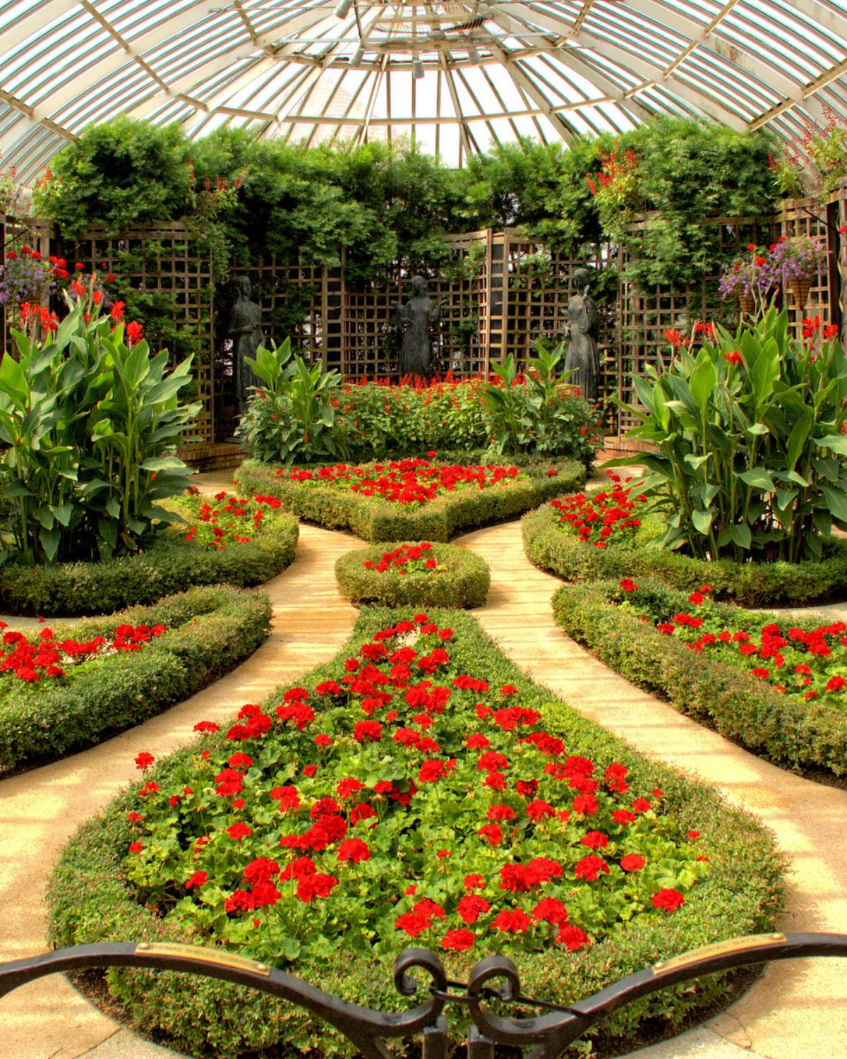 Symmetrical formal garden with red flower beds edged in low hedges, winding sandy paths, and tall green plants and a lattice structure in the background.