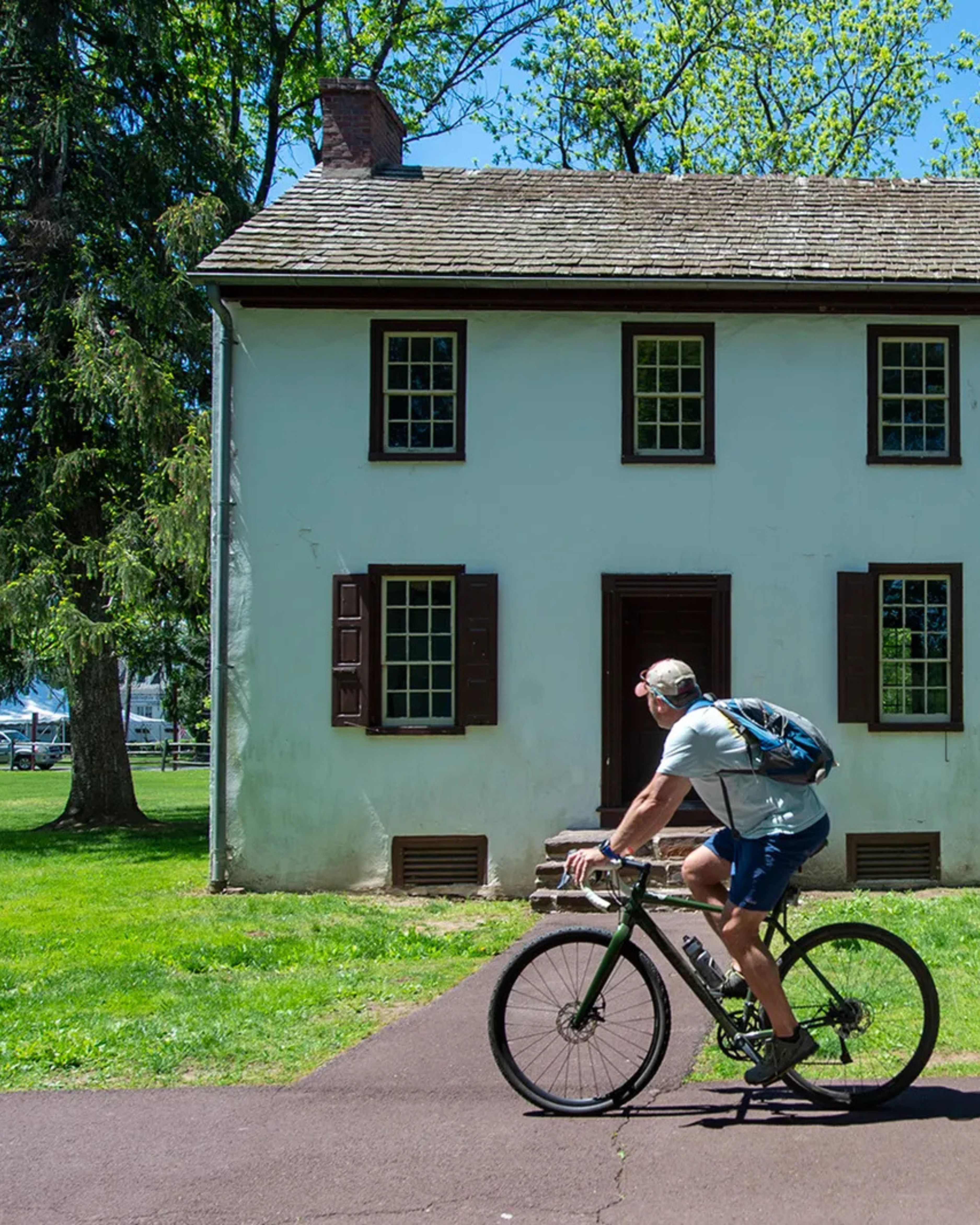 Person riding a bicycle past a historic white colonial-style house at Washington Crossing Historic Park on a sunny day, with green trees and lawn surrounding the building.