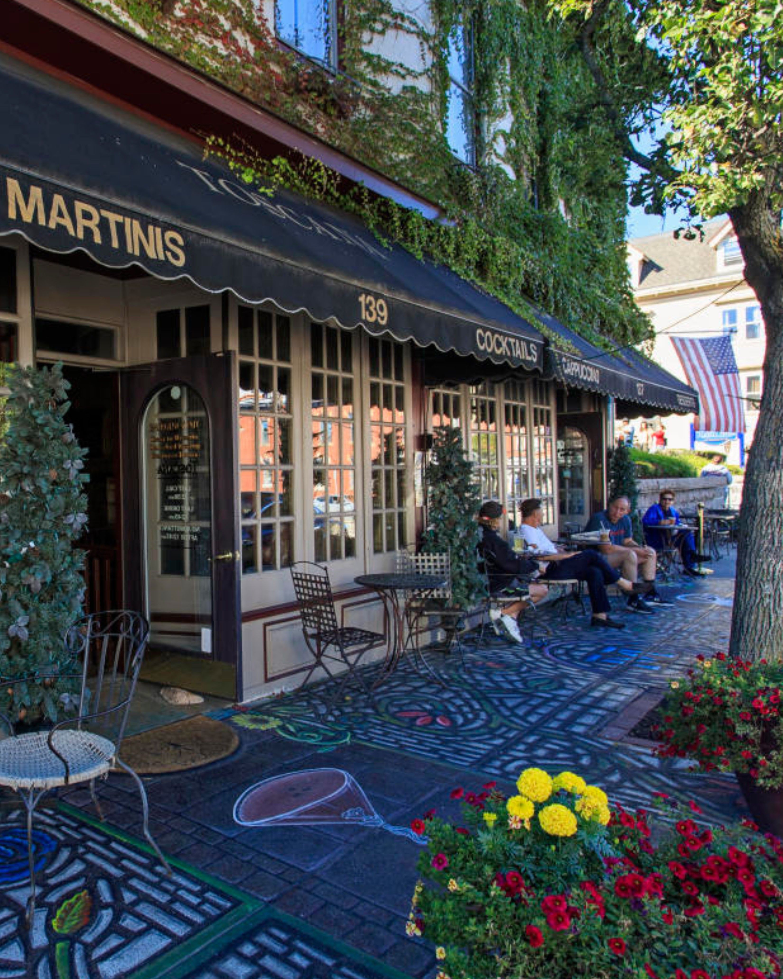 A lively view of Main Street Café in East Greenwich, Rhode Island, featuring cozy outdoor seating under black awnings, ivy-covered storefronts, colorful chalk art along the sidewalk, and guests enjoying drinks and conversation on a sunny day.