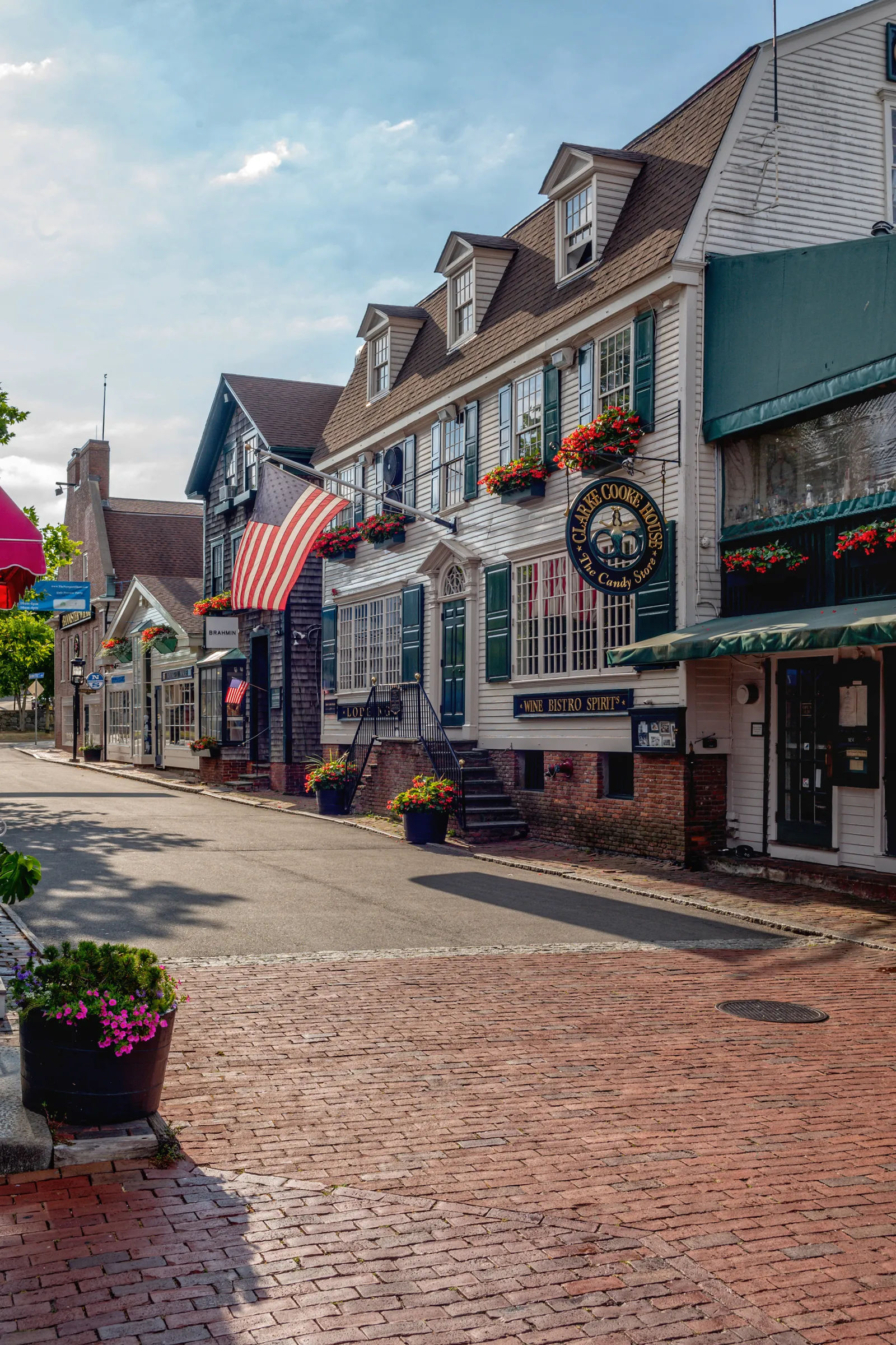 A charming view of Thames Street in downtown Newport, Rhode Island, featuring historic storefronts, brick sidewalks, colorful flower boxes, and classic New England architecture.
