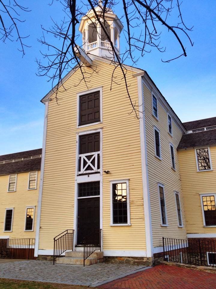 The historic Slater Mill complex in Pawtucket, Rhode Island, featuring the iconic yellow mill building with its cupola, colonial-style architecture, and brick walkways at sunset.