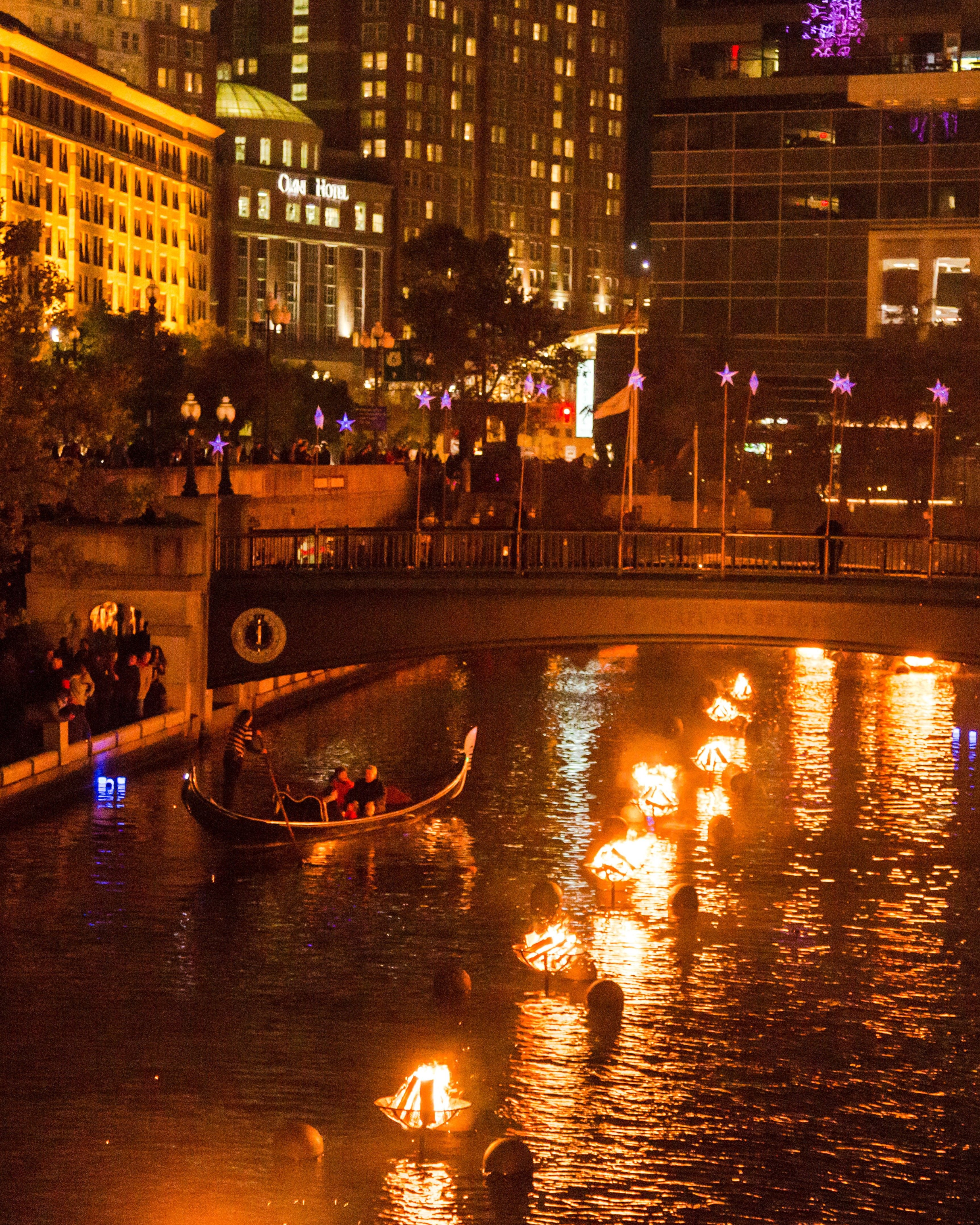 A nighttime view of WaterFire in Providence, Rhode Island, with glowing bonfires floating along the river, a gondola passing under a lit bridge, and crowds gathered along the waterfront surrounded by illuminated city buildings.