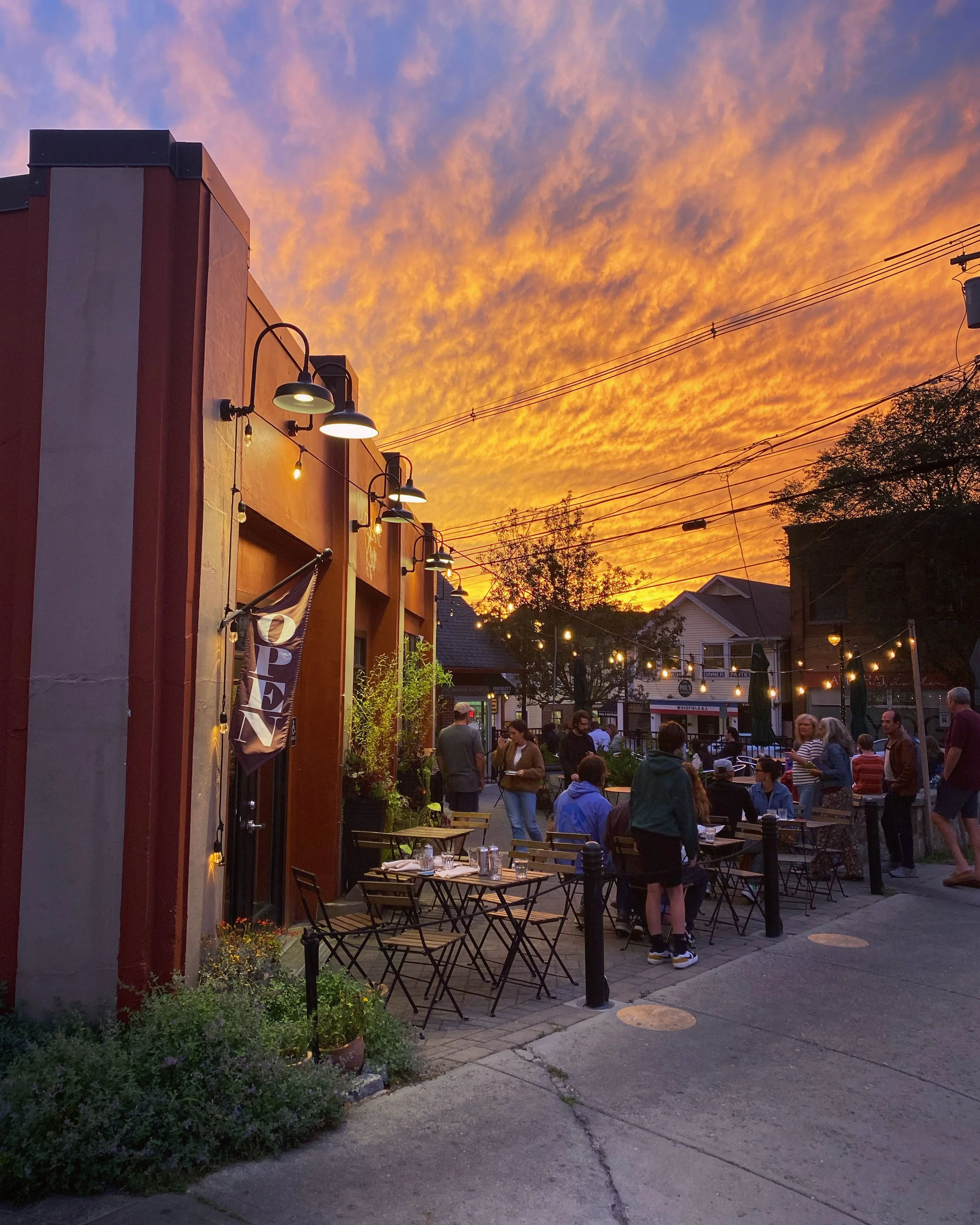 An outdoor dining area in downtown Wakefield, Rhode Island, with restaurant patio seating, string lights, local visitors, and a dramatic sunset illuminating the evening sky.