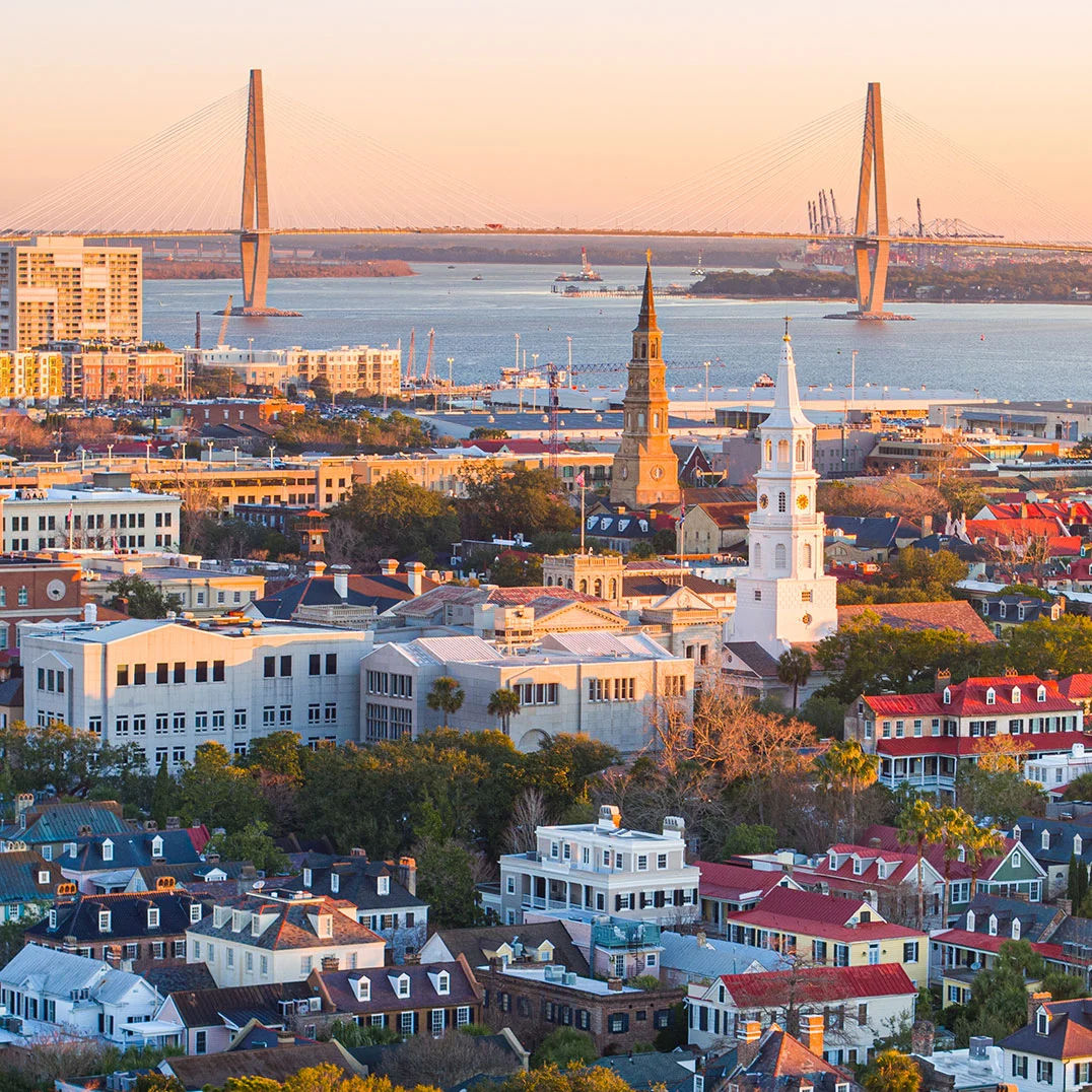 Aerial sunset view of Charleston, SC, featuring historic church steeples, colorful rooftops, and the Arthur Ravenel Jr. Bridge spanning the Cooper River in the background.