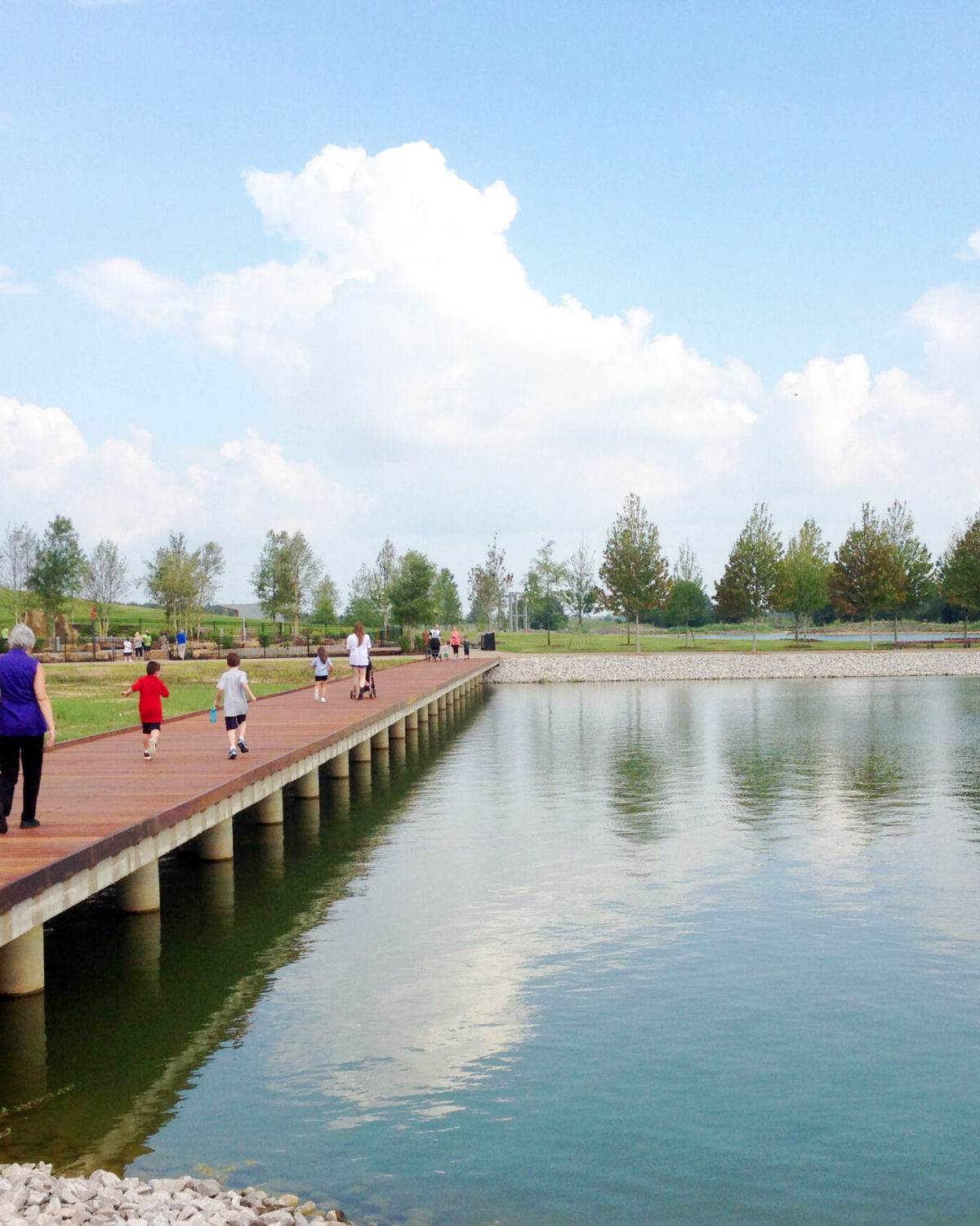 Families walking and jogging along a scenic lakeside boardwalk trail on a sunny day in Cordova, Tennessee