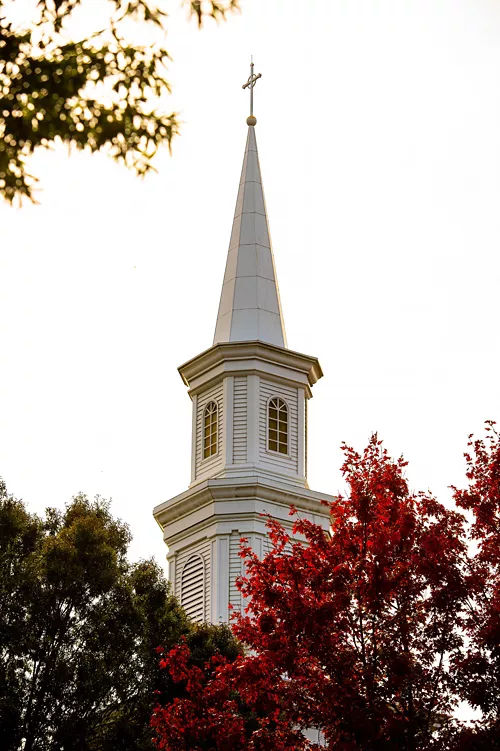 Tall white church steeple with a pointed spire, cross, and arched bell tower windows rising above red autumn leaves and surrounding trees in Germantown, Tennessee. The traditional church architecture stands against a bright, pale sky.