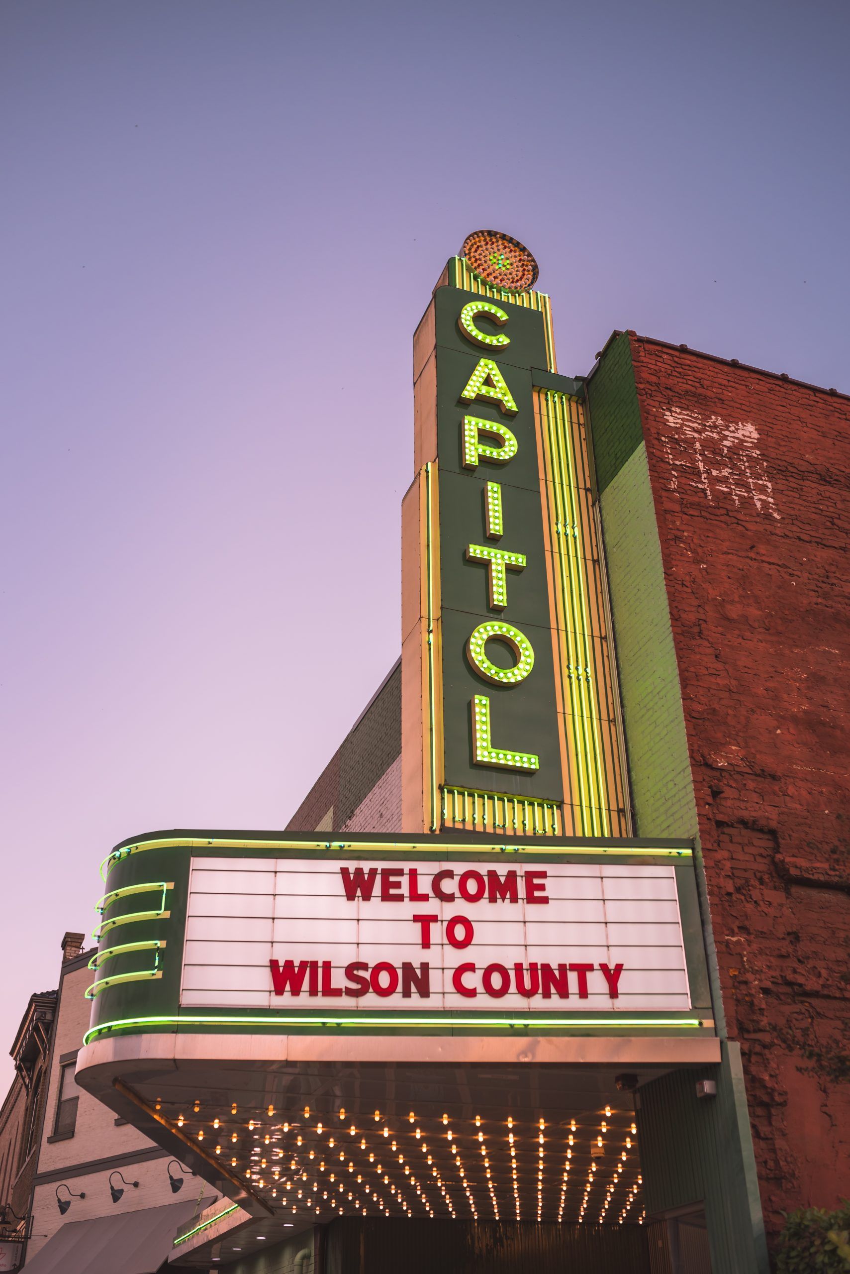 Historic Capitol Theatre in downtown Lebanon, Tennessee, featuring a tall illuminated green vertical sign reading "CAPITOL" and a vintage marquee displaying "WELCOME TO WILSON COUNTY." The theater's brick exterior and glowing marquee lights stand beneath a purple evening sky.