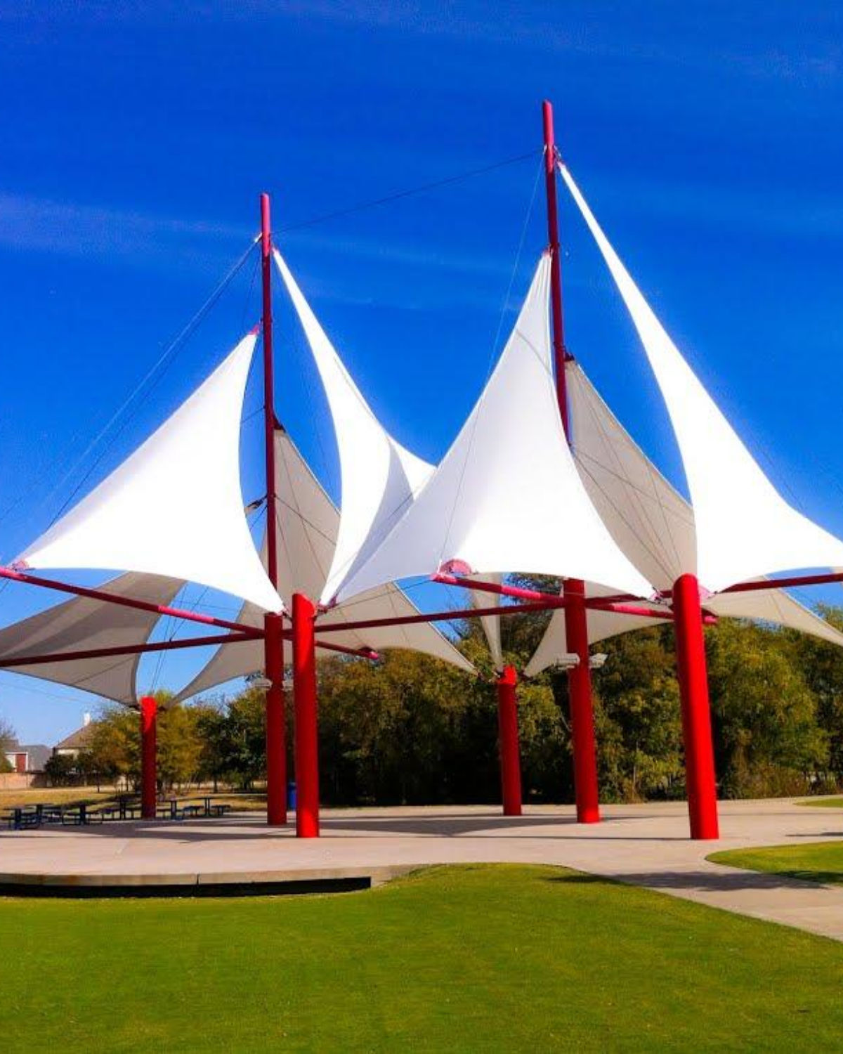 White tensile shade sails stretched over red metal support poles against a bright blue sky, with trees visible below.