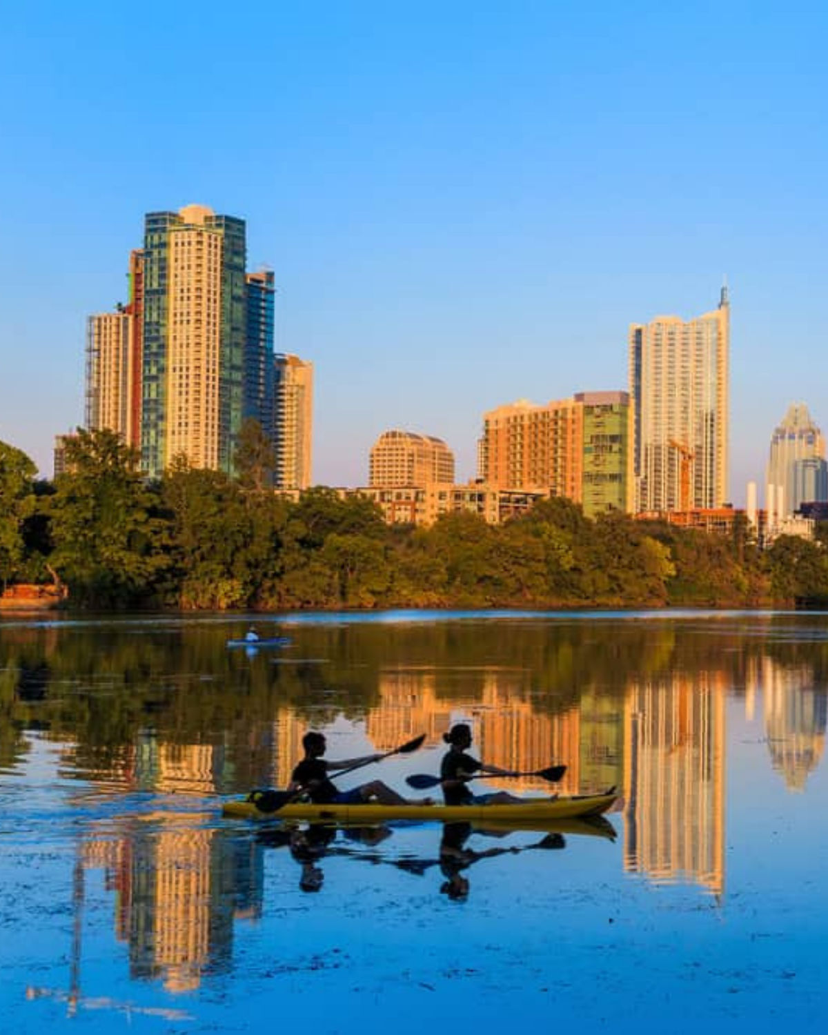Evening view of Austin, Texas, with two people paddling a kayak on a calm river in the foreground and the downtown skyline glowing in golden light, reflected in the water beneath a clear blue sky.