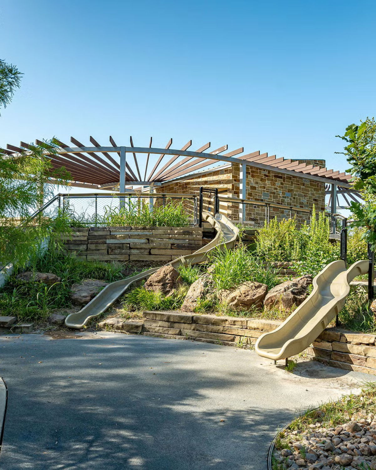 Playground built into a stone-walled hillside with metal slides, a wooden pergola-style shade structure above, and surrounding native plants.