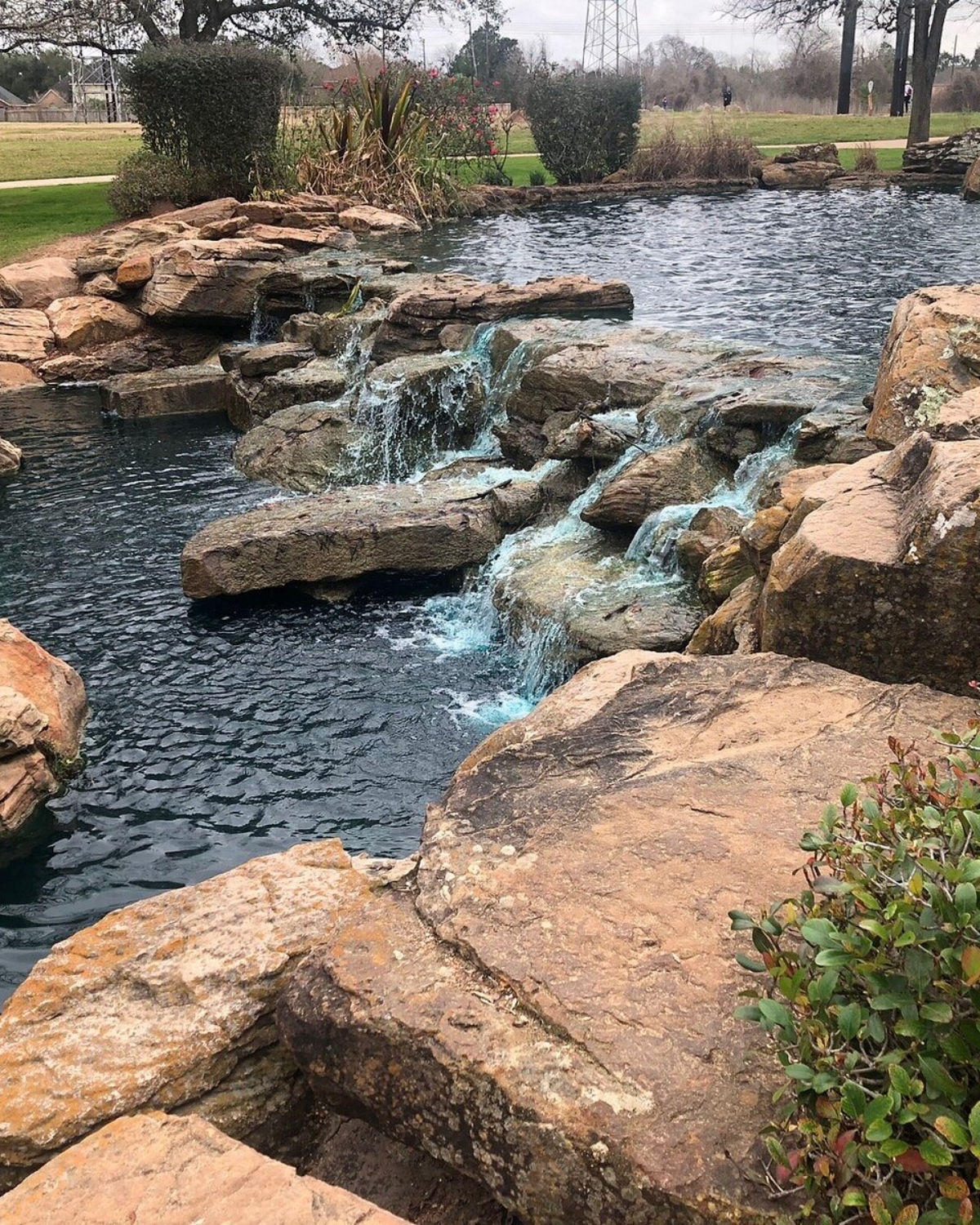 Small waterfall cascading over large tan and orange rocks into a clear pool, with greenery along the right edge.