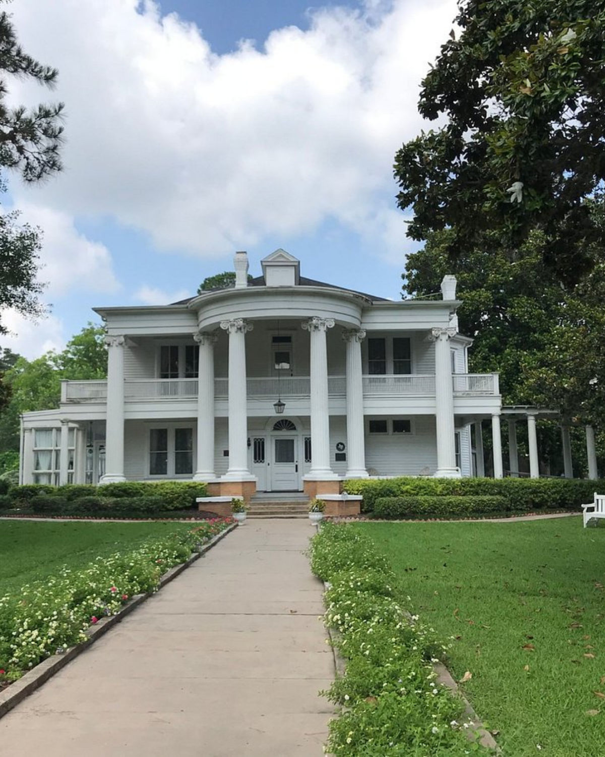 Front view of the Fort Bend Museum in Richmond, Texas, showing a grand white historic mansion with tall Corinthian columns, a curved two-story portico, and a flower-lined walkway leading through the manicured lawn to the entrance.