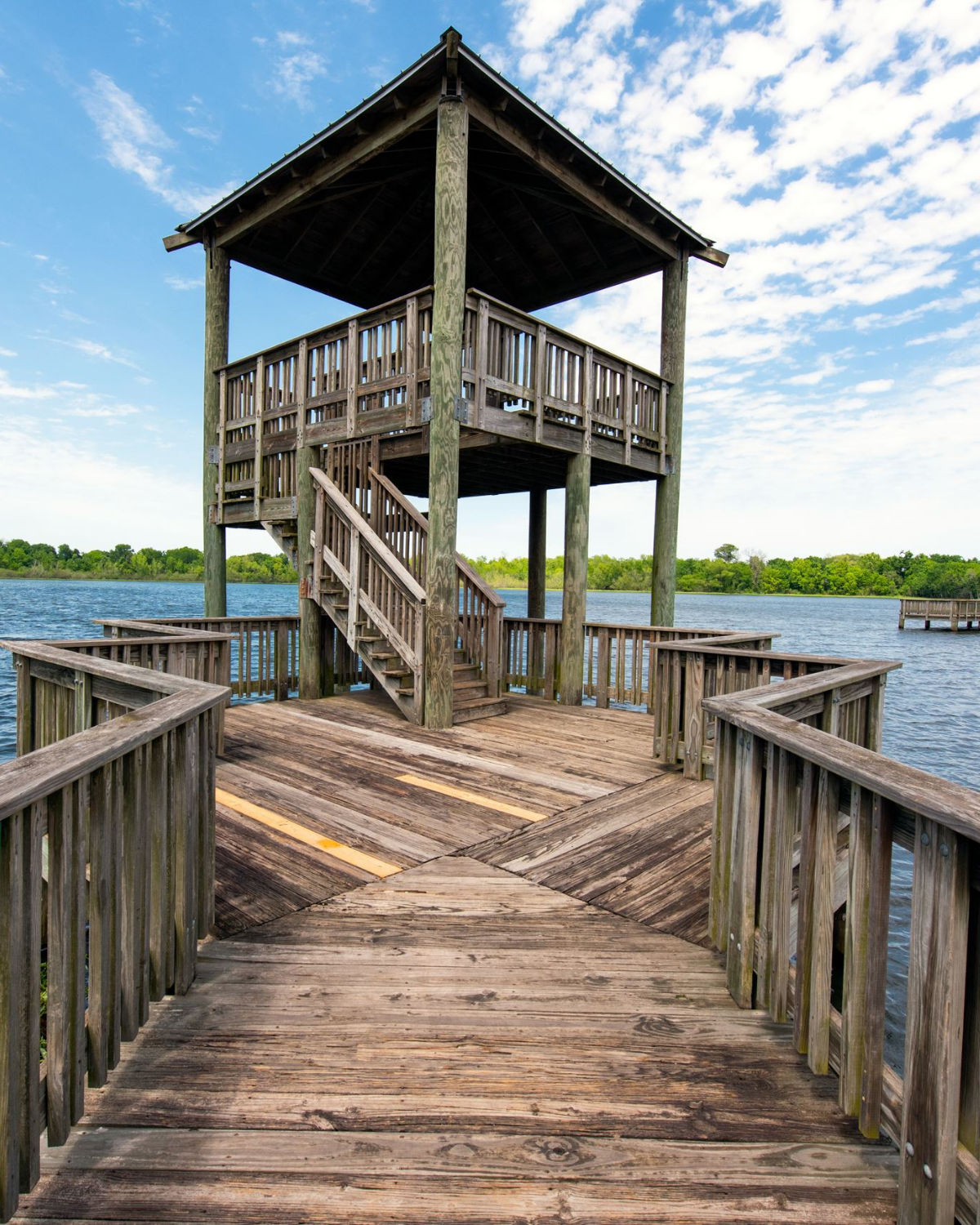 A quiet wetland scene at Cullinan Park in Sugar Land, Texas, featuring a rustic wooden observation tower and boardwalk rising above still water dotted with lily pads and backed by a treeline under a soft evening sky.