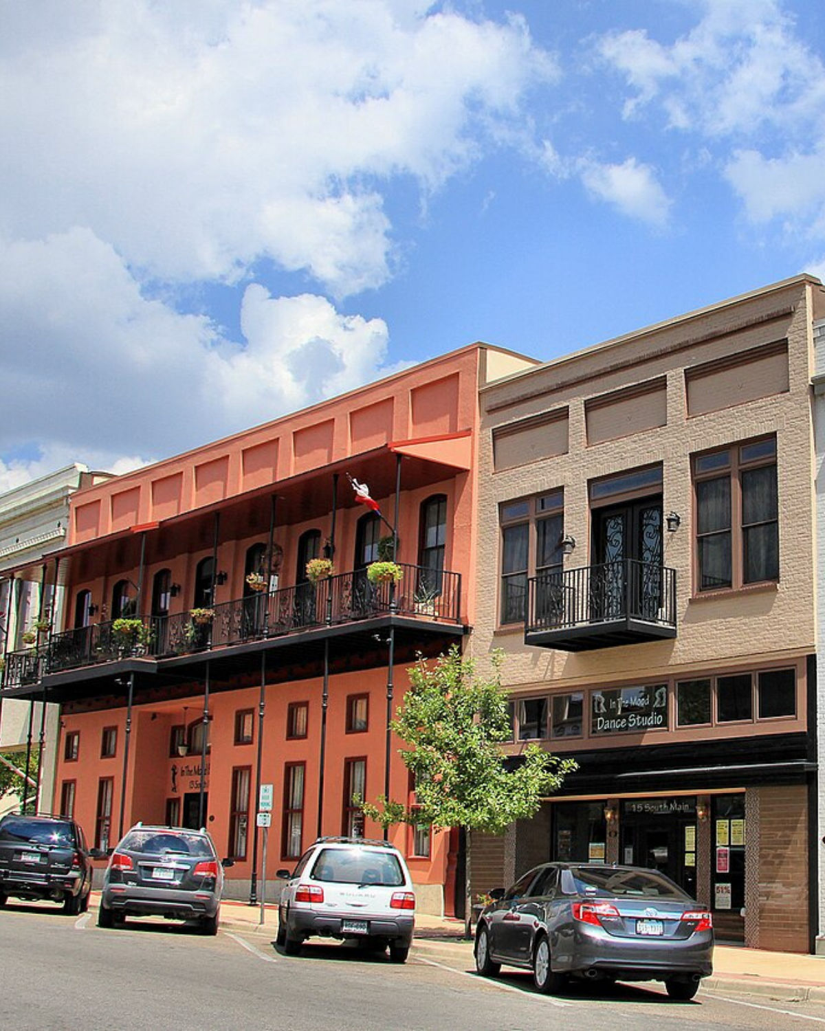 Colorful historic storefronts along downtown Temple, Texas, with warm-toned facades, second-story iron balconies, and parked cars lining the street under a bright, partly cloudy sky.