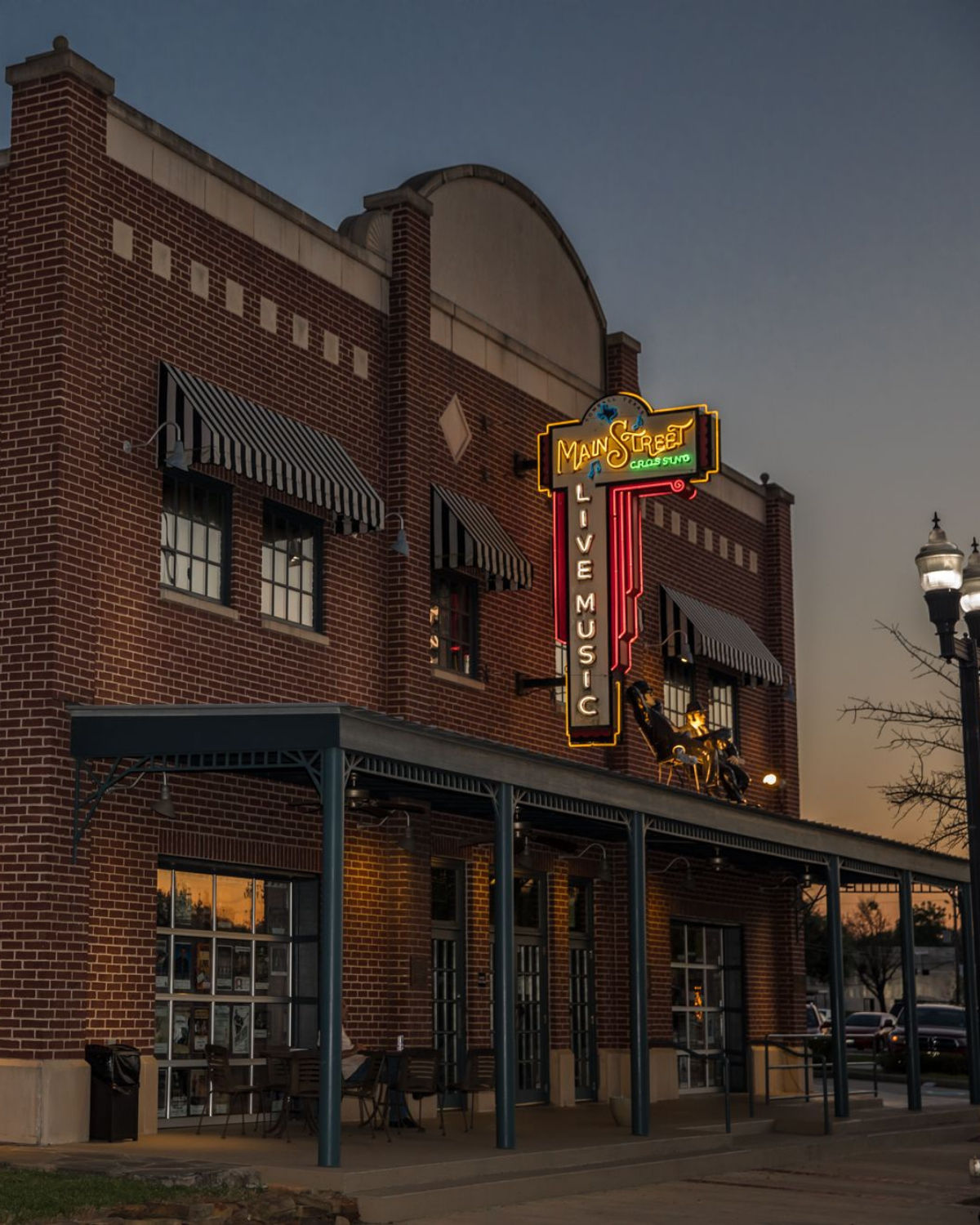 Brick-front Main Street Crossing music venue in Tomball, Texas, at dusk, with striped awnings, a covered sidewalk patio, and a glowing vintage-style neon sign advertising live music against the evening sky.