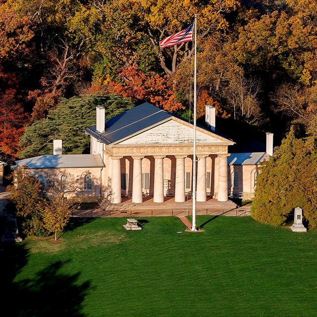 The Arlington House in Arlington, Virginia, with its iconic columned façade, American flag in front, and colorful fall foliage surrounding the historic estate.