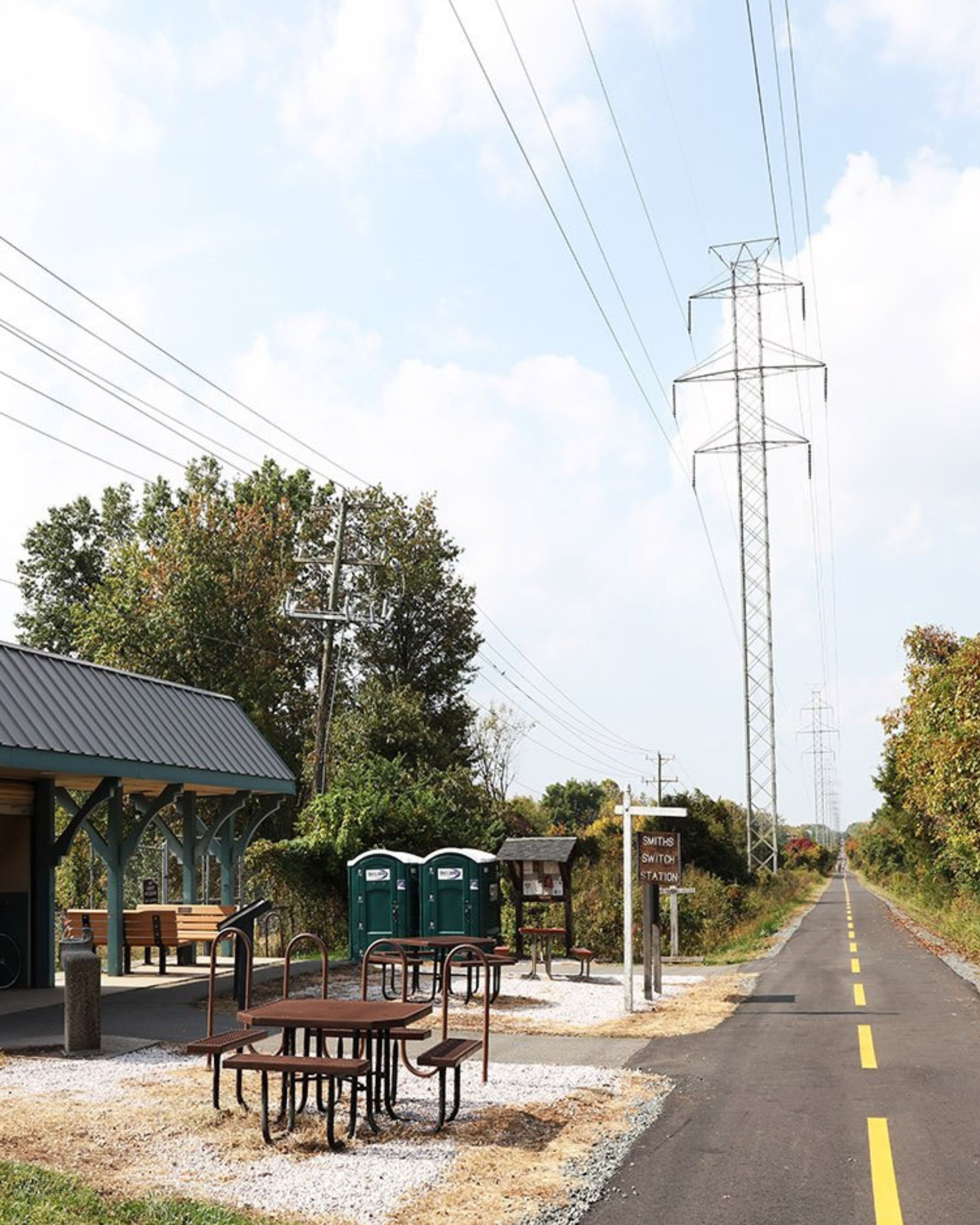 A paved stretch of the Washington & Old Dominion Trail in Ashburn, Virginia, featuring a shaded rest area with picnic tables, benches, and a small shelter, with power lines running overhead and trees lining the path on both sides.