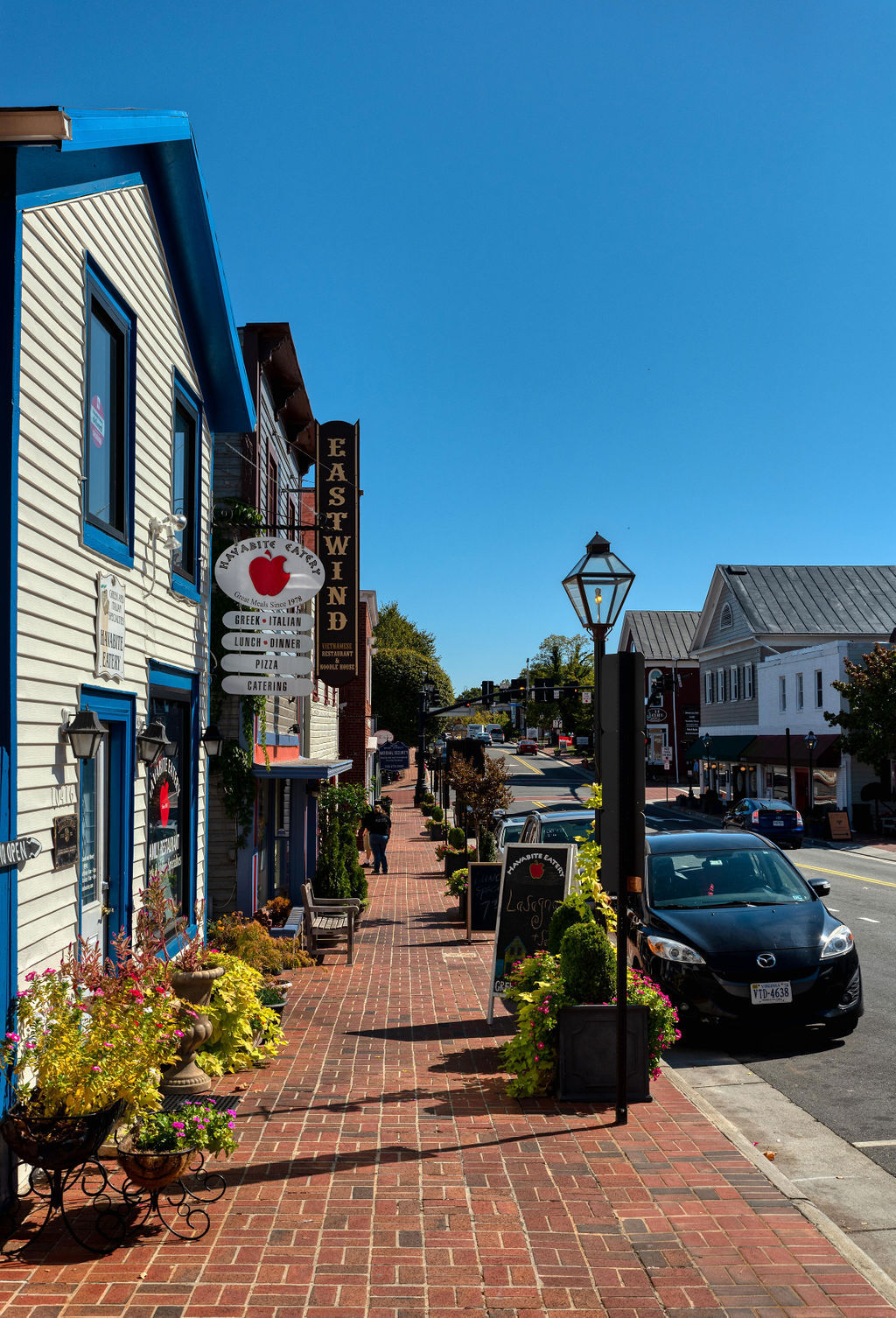 A charming Fairfax street lined with brick sidewalks, local shops, and restaurants, featuring colorful storefronts, outdoor seating, and classic lantern-style streetlights under a clear blue sky.
