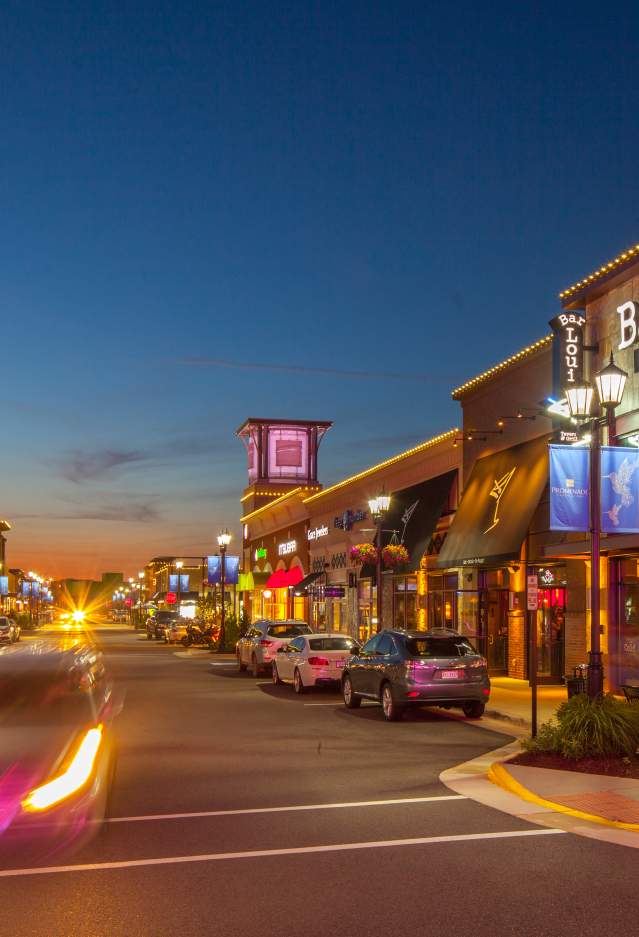 A lively evening view of Virginia Gateway in Gainesville, Virginia, featuring warmly lit storefronts, popular restaurants like Bar Louie, and a bustling street scene with passing cars and pedestrians enjoying the vibrant nightlife atmosphere.