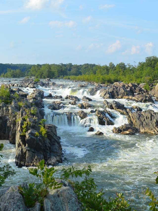 Great Falls Park in Great Falls, VA, featuring powerful waterfalls and rushing rapids cascading through rugged rock formations along the Potomac River under a clear blue sky.