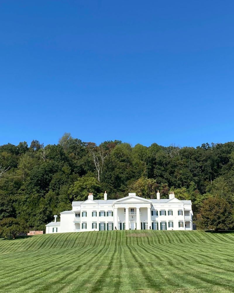 Exterior view of Morven Park in Leesburg, Virginia, featuring the historic white mansion, expansive manicured lawn, and wooded hillside backdrop beneath a clear blue sky.