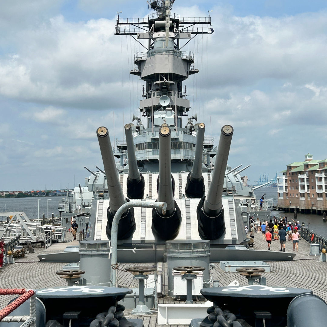Front-facing view of the USS Wisconsin at Nauticus in Norfolk, VA, featuring large naval artillery guns on deck with visitors walking nearby and waterfront views in the background.