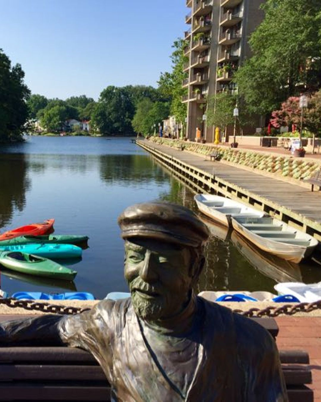 A peaceful view of Lake Anne in Reston, Virginia, featuring colorful kayaks and paddle boats along the water’s edge, with a bronze statue in the foreground and a scenic boardwalk lined with trees and buildings in the background.