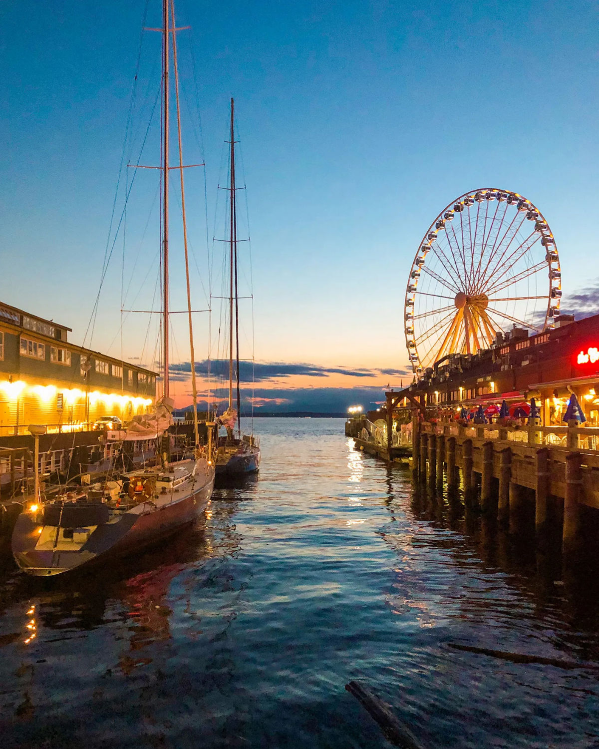 Twilight view of Seattle’s waterfront with sailboats moored along the pier and the illuminated Seattle Great Wheel rising above restaurants and shops, reflected in the calm waters of Elliott Bay beneath a glowing sunset sky.