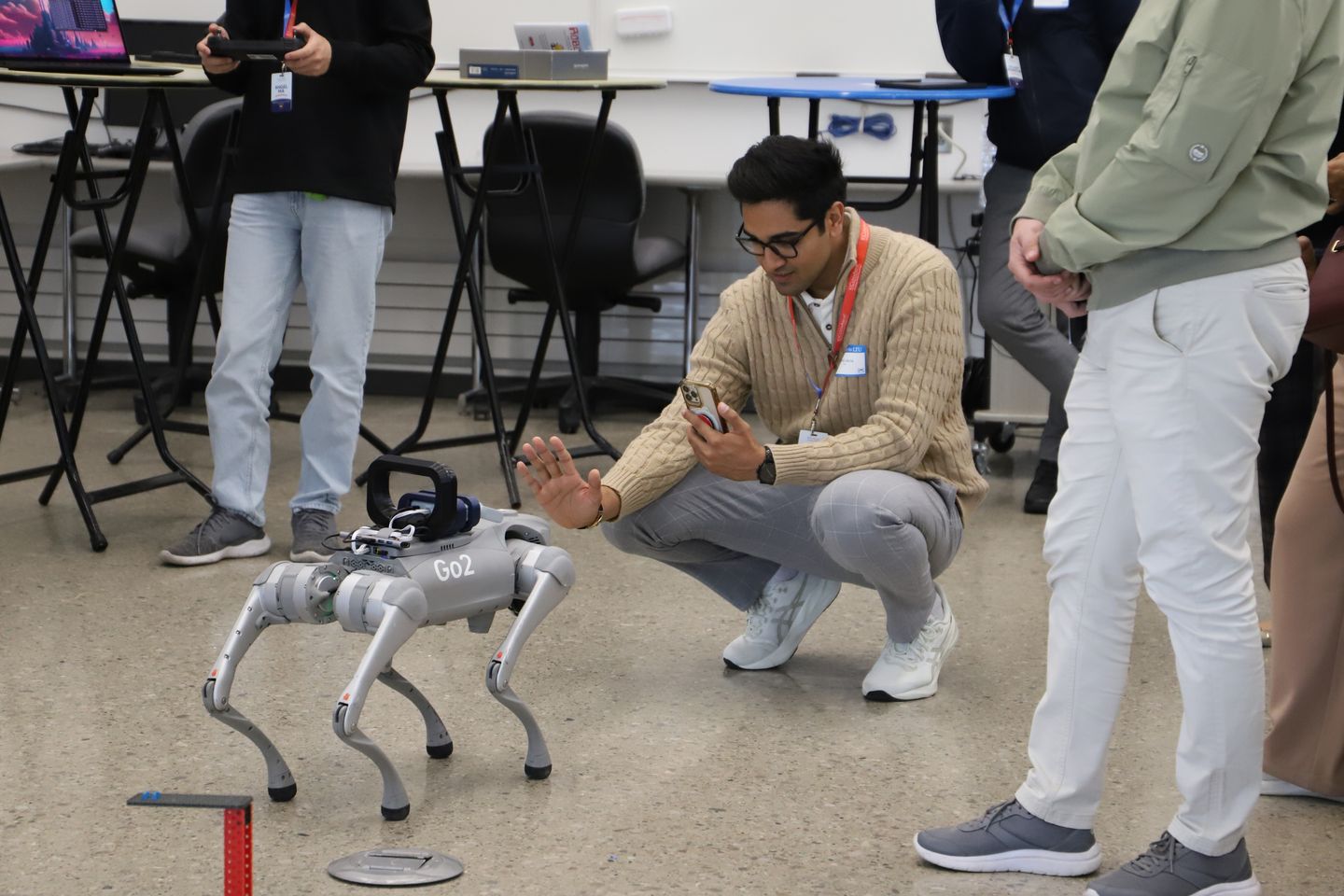 An international student counselor crouches while using a smartphone to interact with a gray quadrupedal robot.