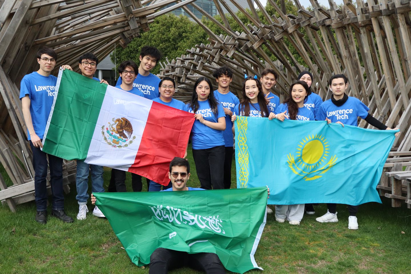 A group of international students wearing Lawrence Tech shirts stand outdoors holding the flags of Mexico, Kazakhstan, and Saudi Arabia in front of a wooden, latticework structure created by LTU's Architecture students.