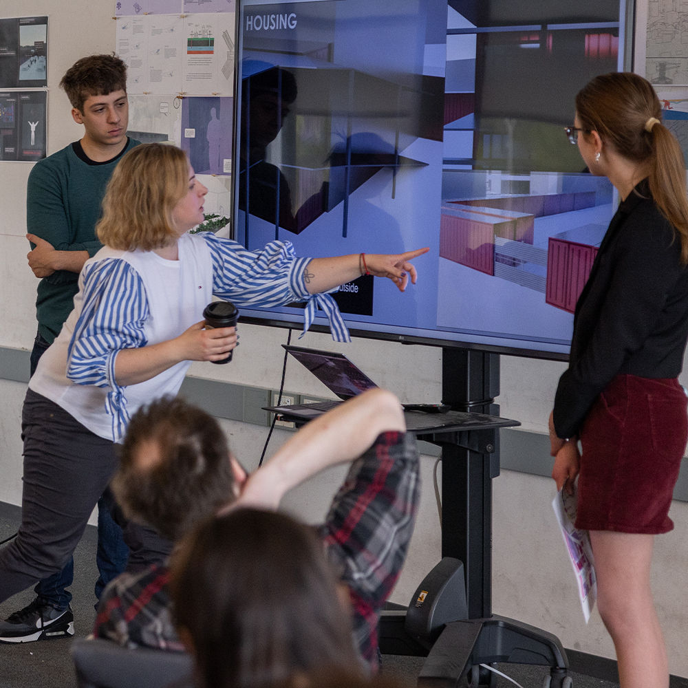 A student presents an architectural rendering on a large monitor to a small group of peers in a classroom setting. A faculty member points to the screen, giving feedback.