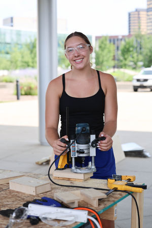 A smiling student wearing safety glasses poses behind a workbench while holding a power router tool on a piece of wood.