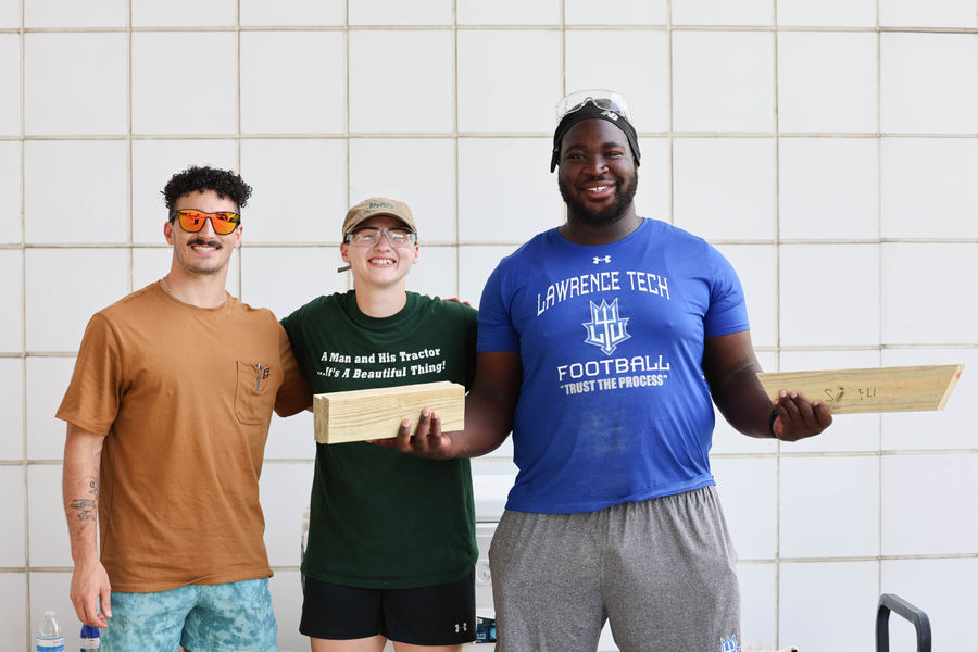 Three smiling students stand side-by-side against a white tiled wall, with two of them holding pieces of cut lumber.