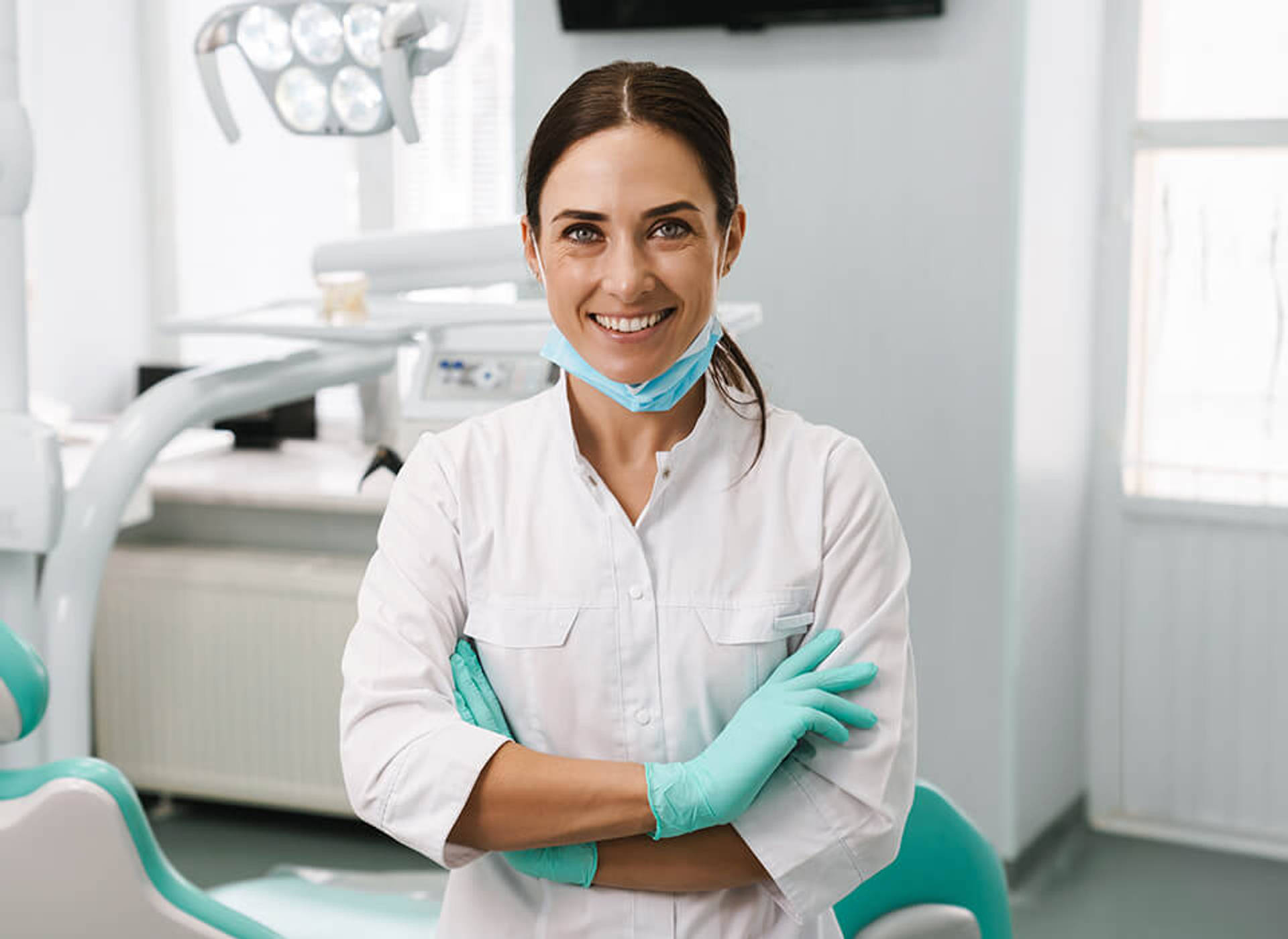 A smiling orthodontic specialist in a white coat with her arms crossed in a treatment room.