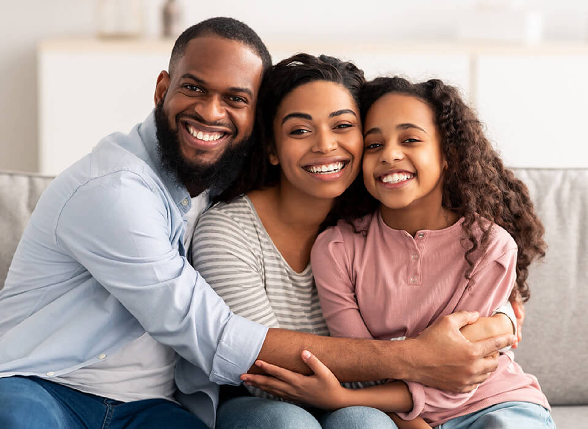 A smiling father, mother, and daughter hugging on a couch.