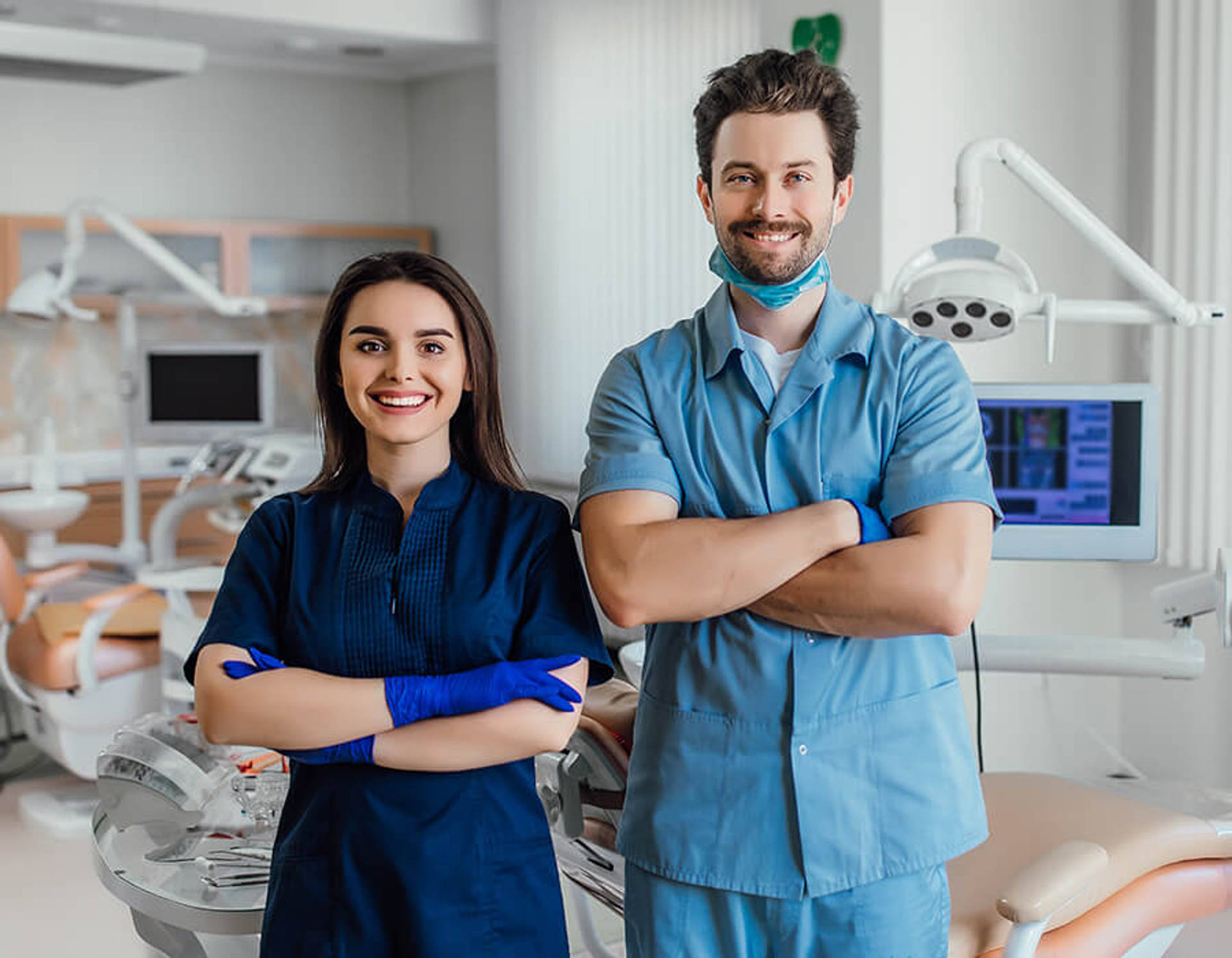 Two smiling orthodontic specialists in scrubs standing side by side with their arms crossed in a treatment room.