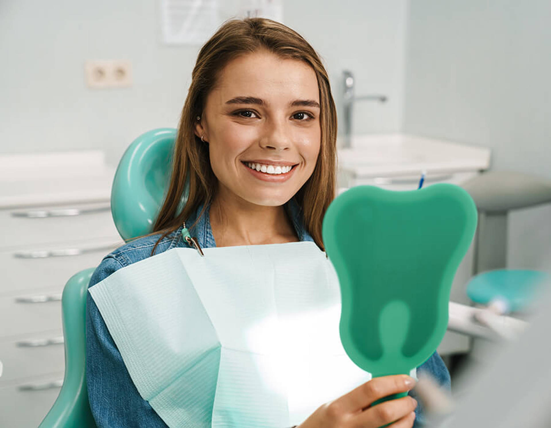 A smiling young woman holding a tooth-shaped mirror while seated in an orthodontic treatment chair.