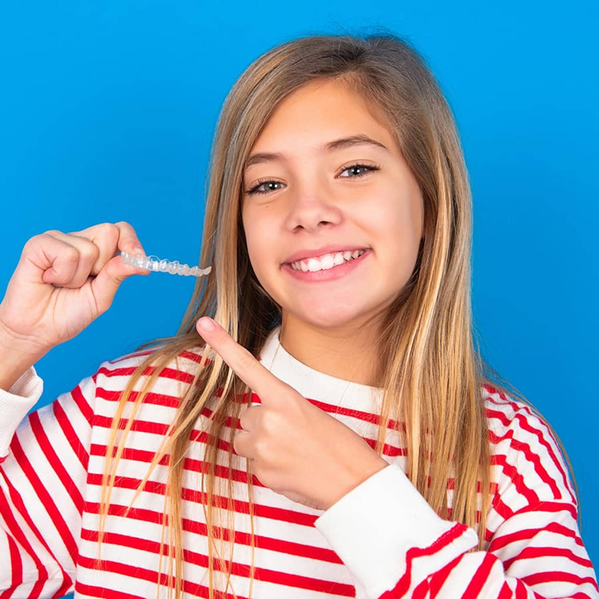 caucasian teen girl wearing striped shirt over blue studio background holding an invisible aligner and pointing at it. Dental healthcare and confidence concept.