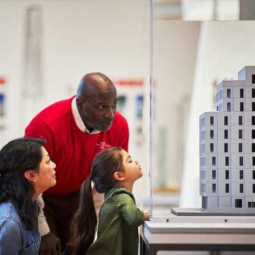 A tall bald man in a red shirt leaning over two people, one an older person with short hair and the other a young person in a green top