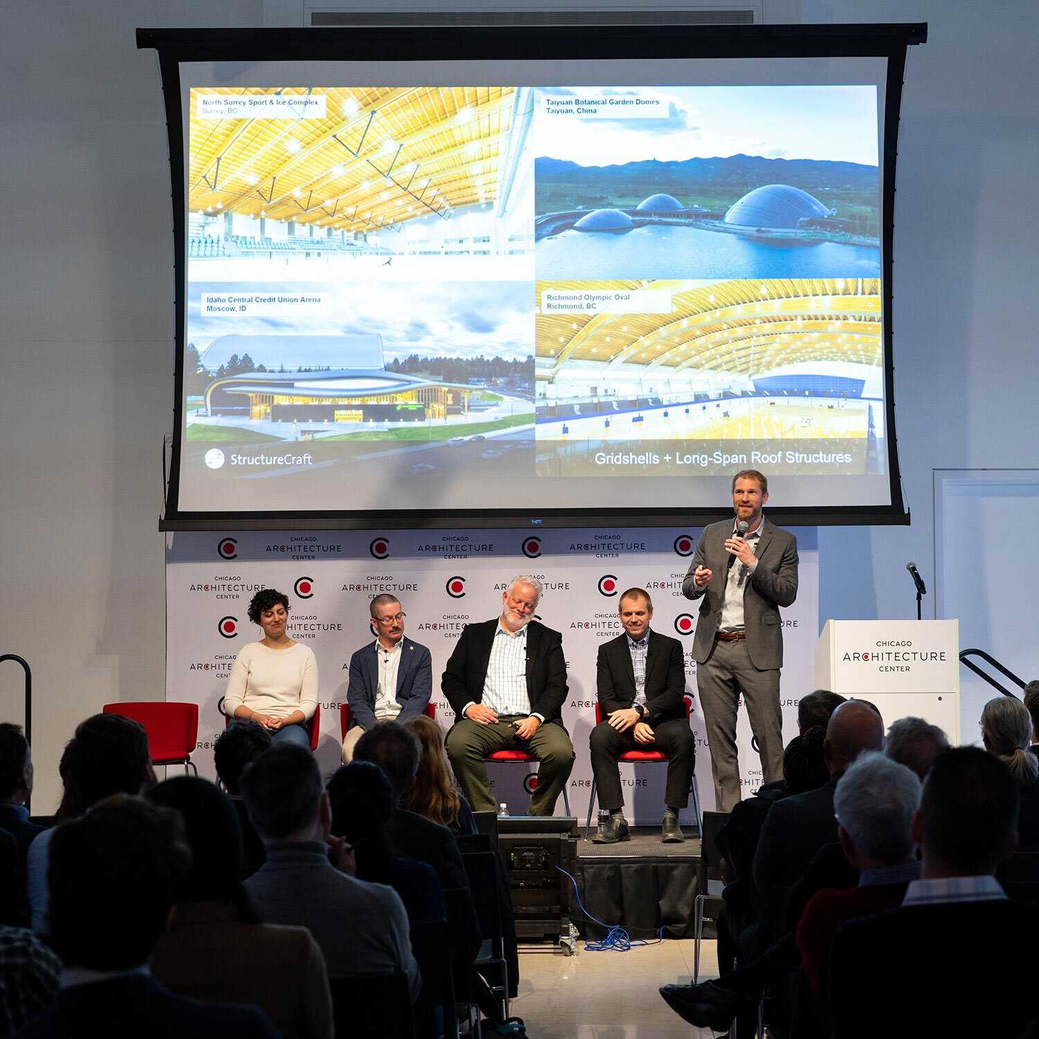 A panel of 4 men and 1 woman in front of a projector screen. The man on the right is standing and holding a microphone. 