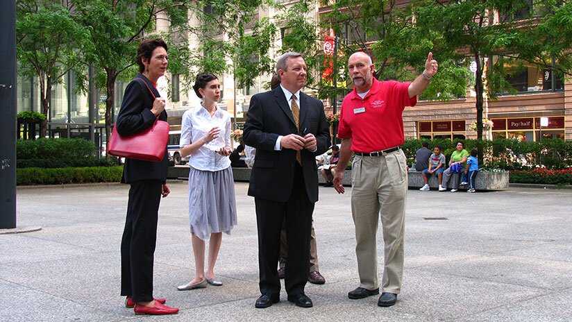 Small group of folks in business attire listening to a docent in a red shirt describing what they are seeing