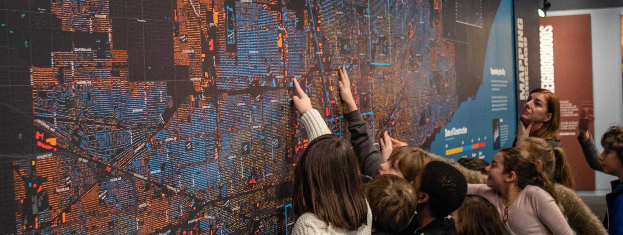 Group of kids pointing on a map wall with teacher looking on.