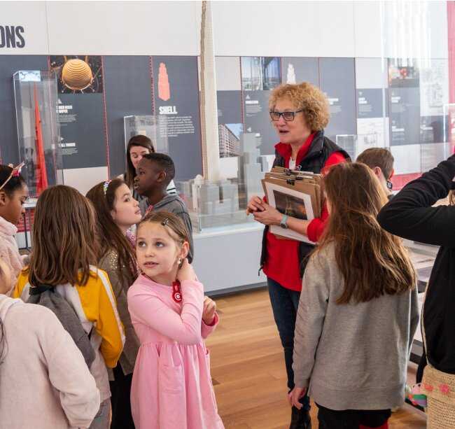 A group of children in a gallery overseen by a person with short hair in red shirt and black vest