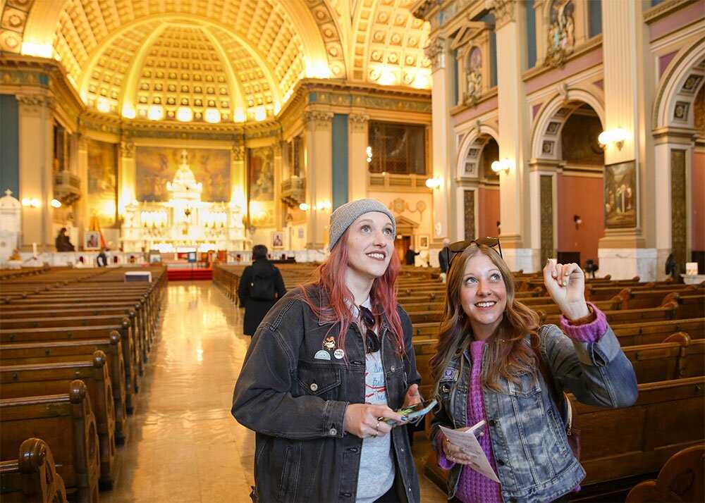 Two young women looking at something intently while they stand inside a beautifully lit church inside