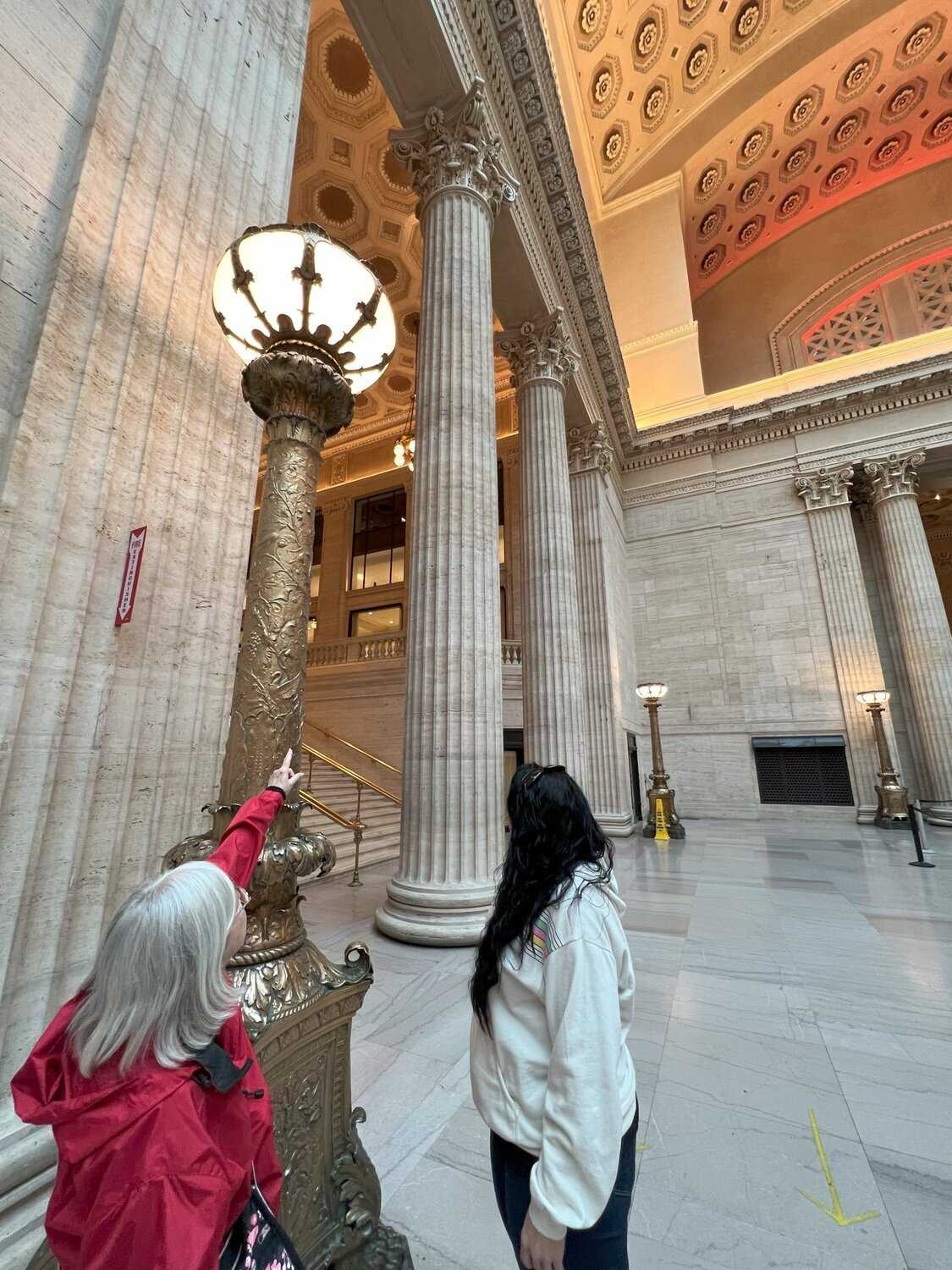 Photo of a docent and tour goer. The docwent is pointing at a golden light post and there are columns and a valuted ceiling in the background. 