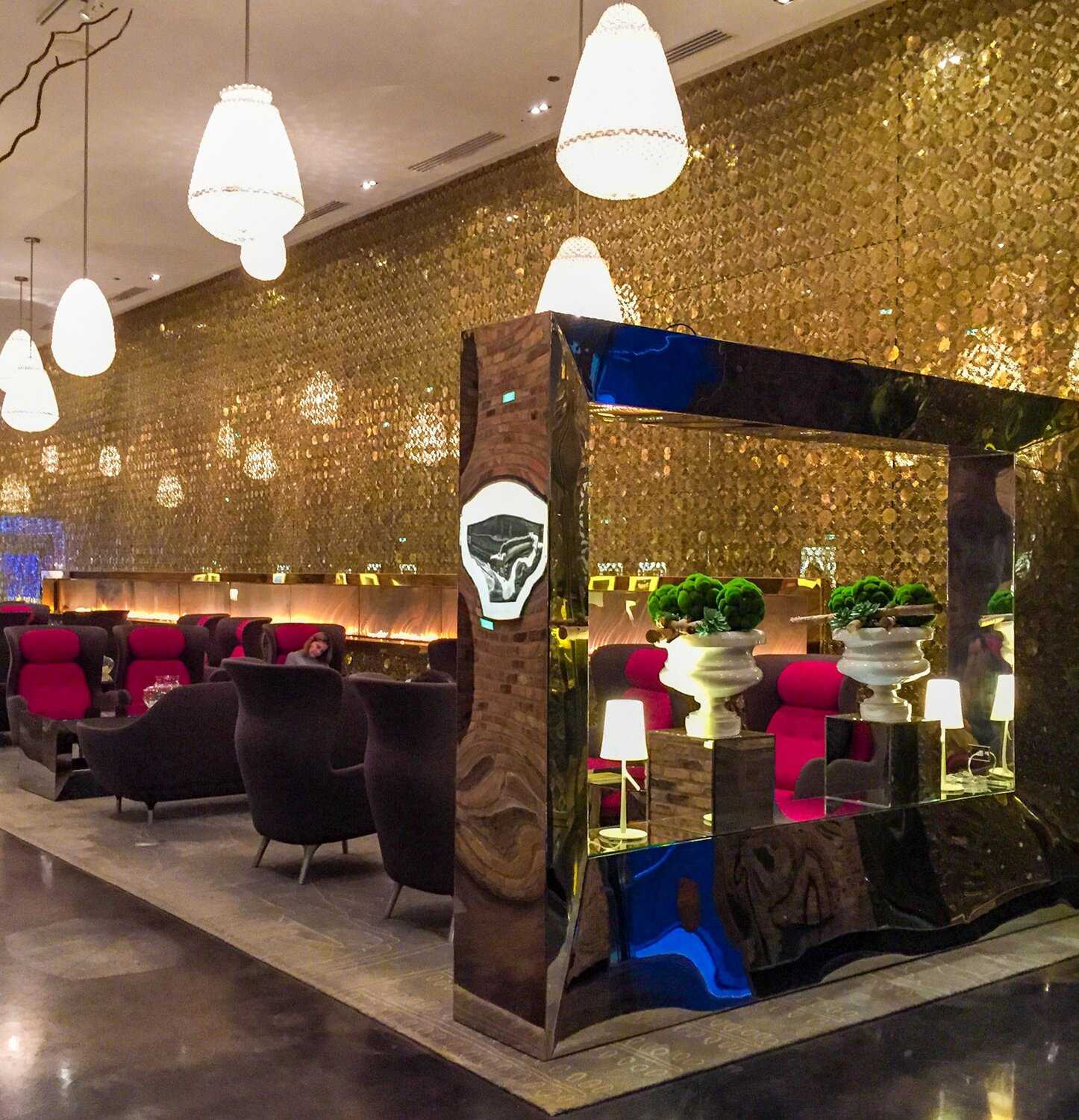 Image of a hotel lobby with purple wingback chairs, pendant lights hanging from the ceiling and a golden wall. 